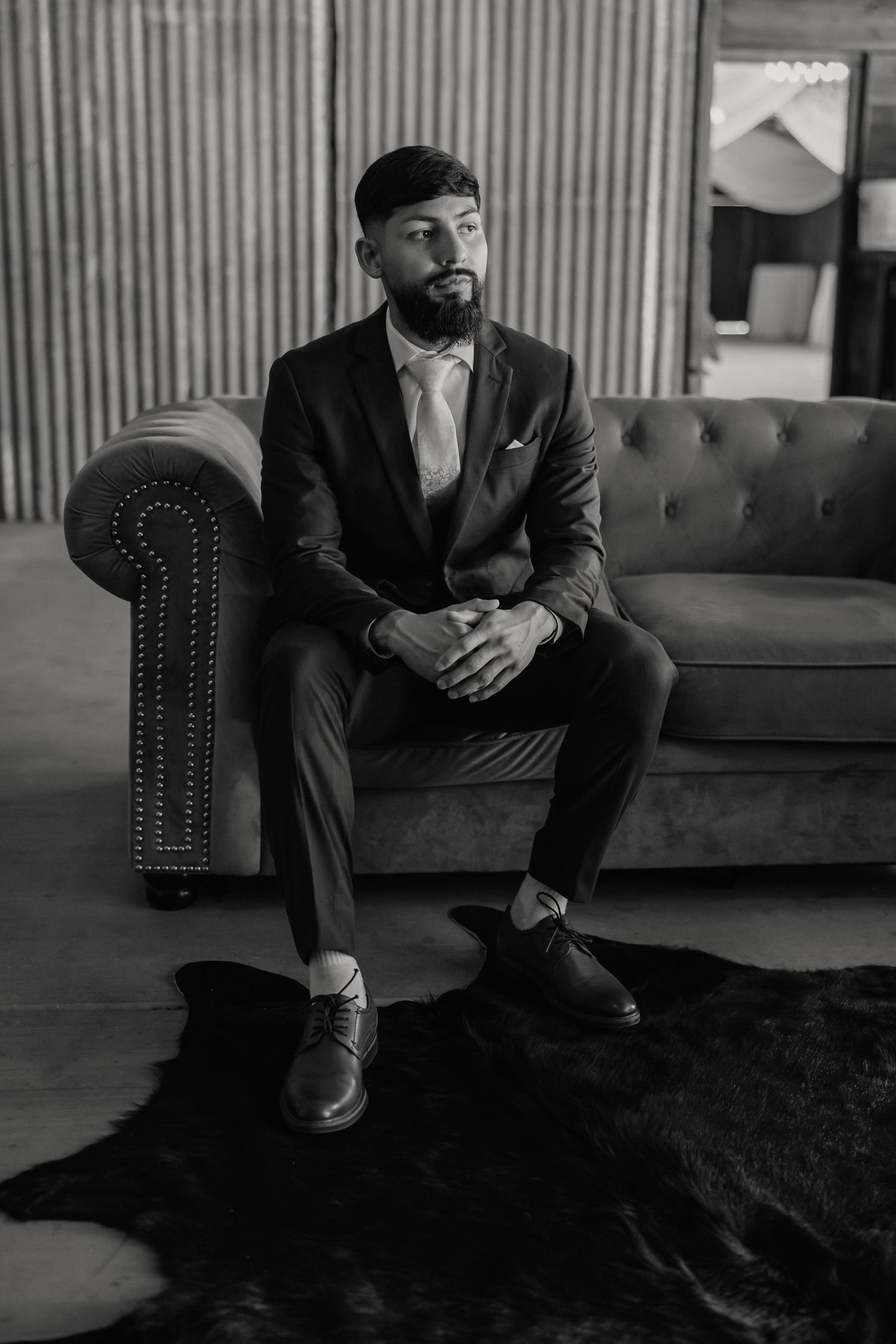 Groom in black suit sits on tufted sofa in black and white portrait photo.