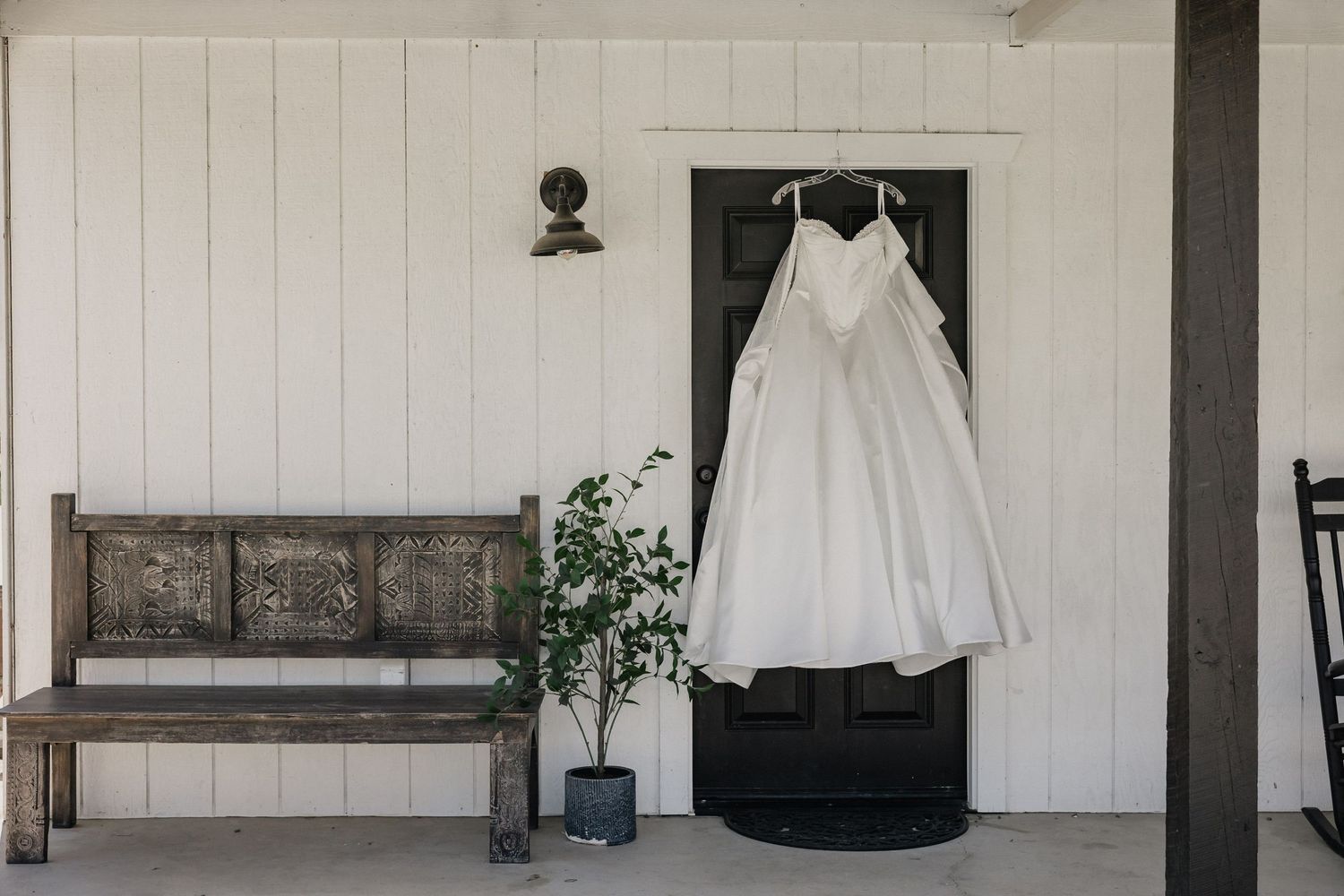 White ball gown wedding dress hanging on door of rustic white farmhouse porch with wooden bench.