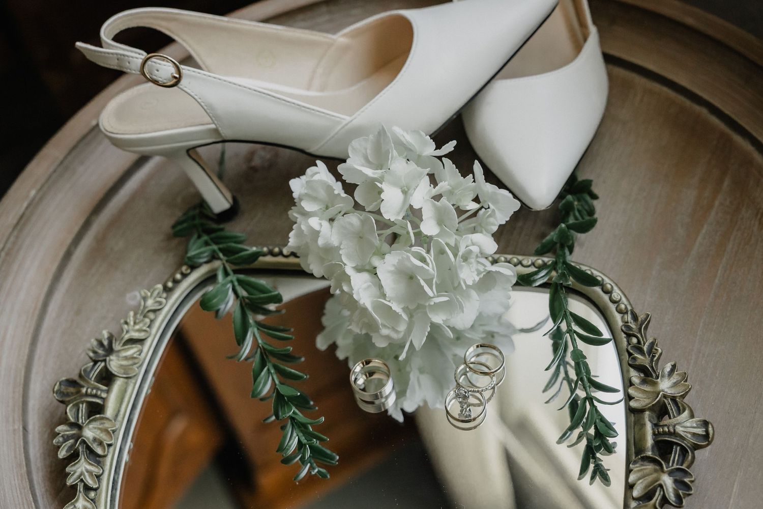 White bridal heels and hydrangea boutonniere displayed on ornate vintage mirror wedding detail.