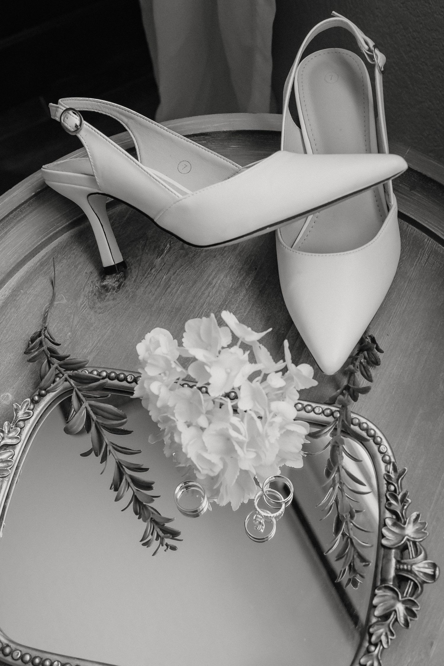 Black and white flat lay of white bridal heels, wedding rings, and delicate flowers on an ornate mirror tray.