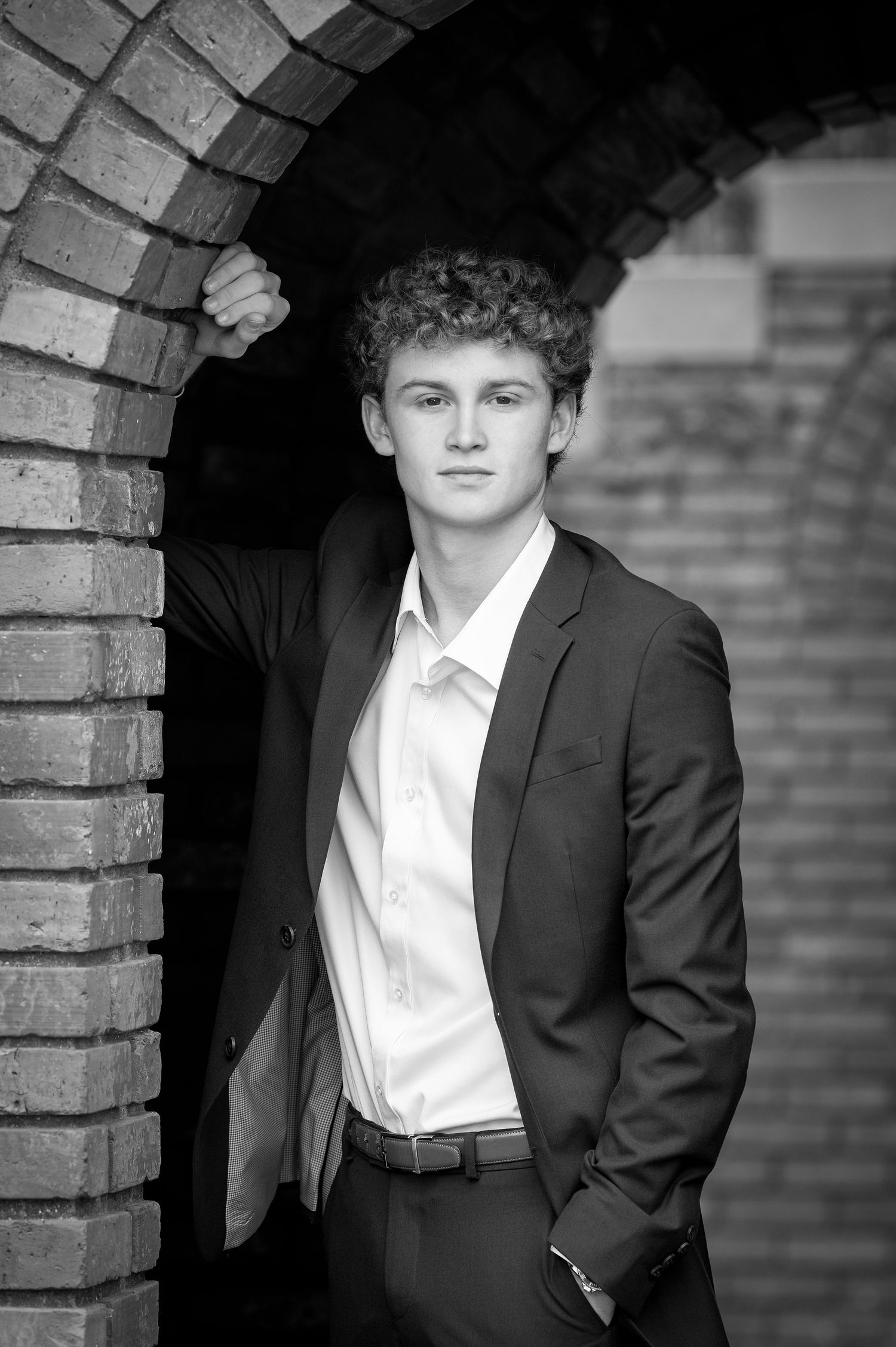 Young man in dark suit and white shirt poses by brick archway in black and white portrait photo.
