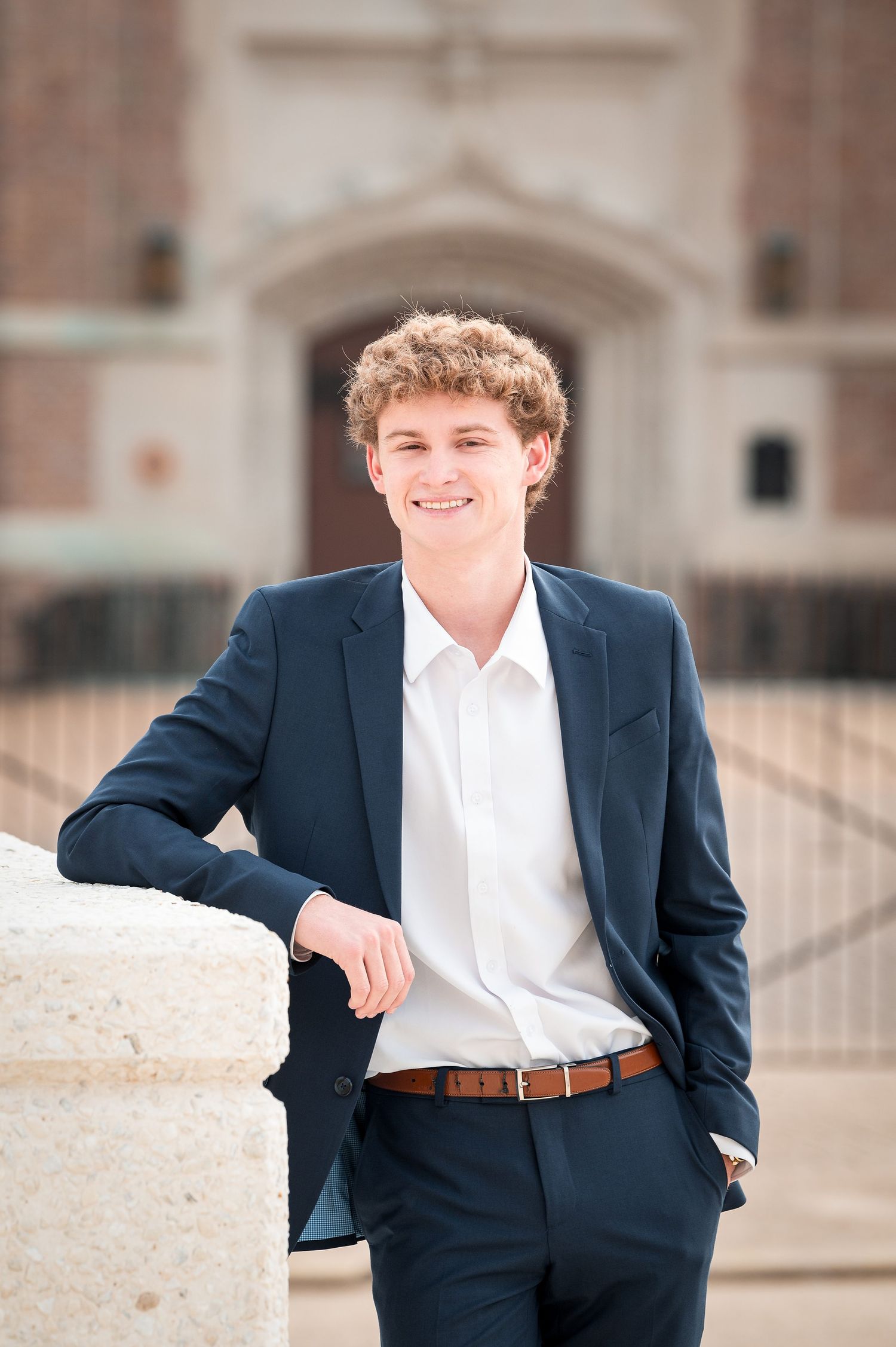Young man in navy suit and white shirt smiling outdoors near brick building.