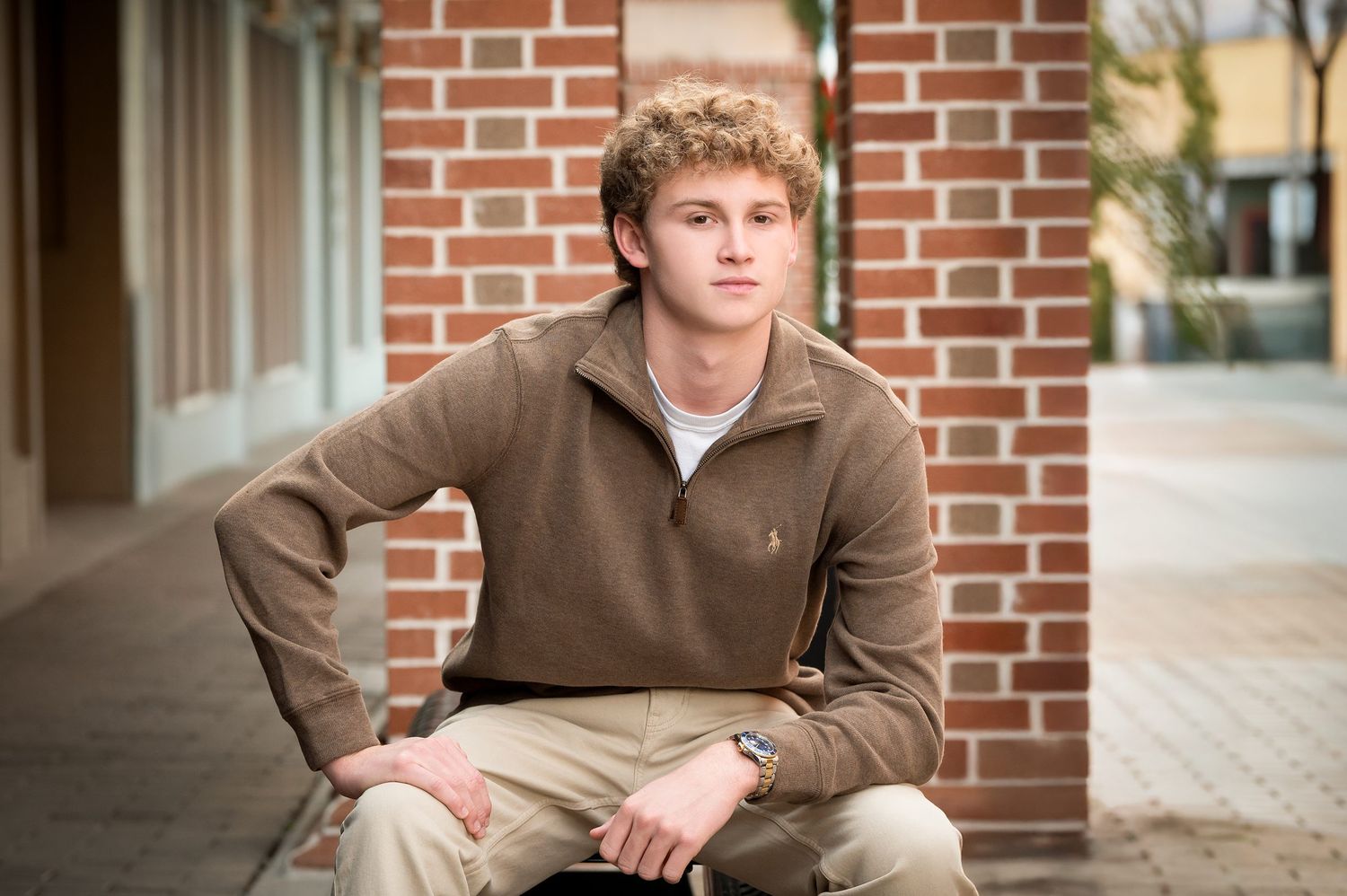 Young man in brown pullover and khakis sitting outdoors near brick wall.