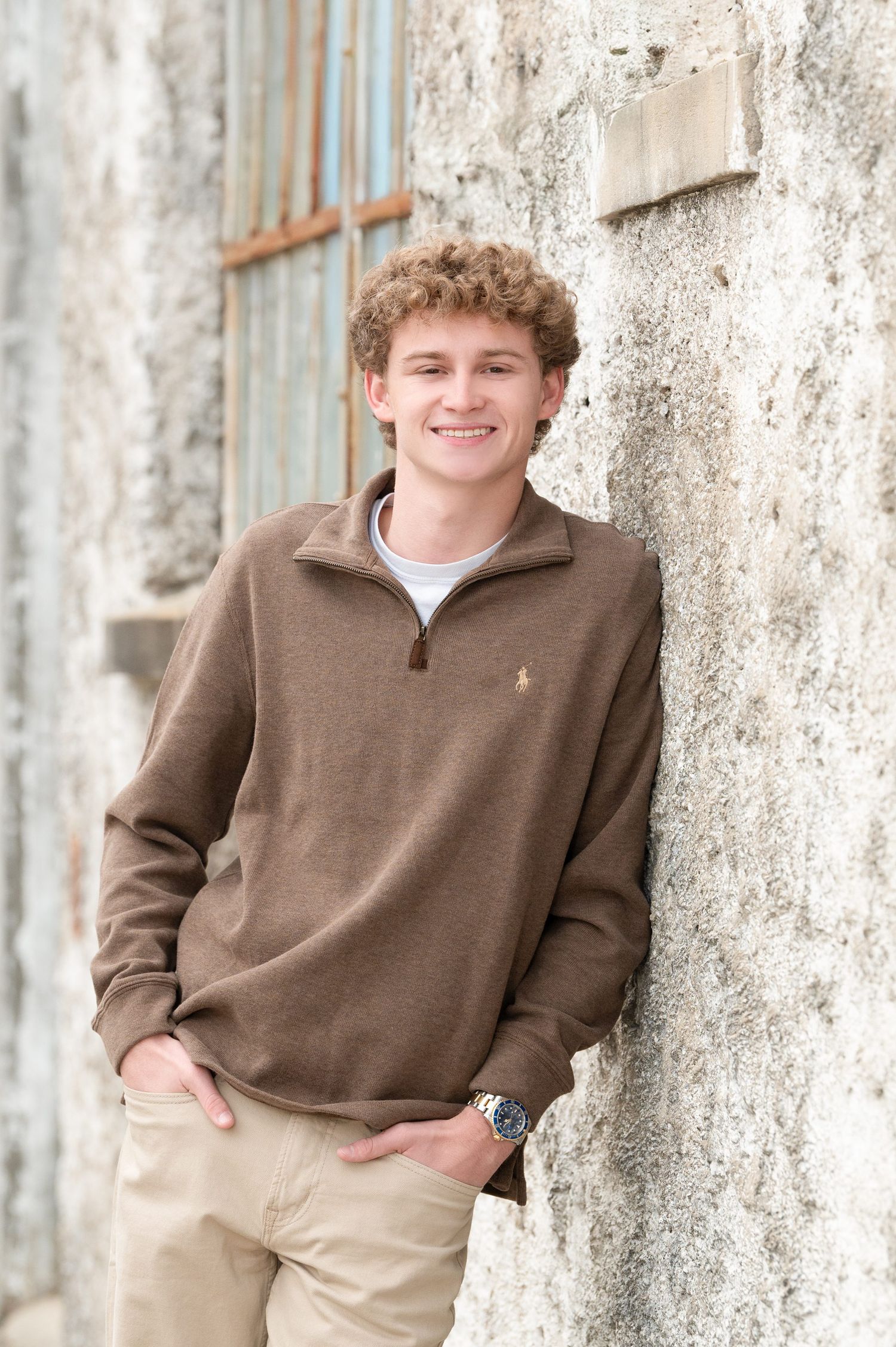 Smiling young man with curly hair leaning against a stone wall wearing a brown Polo quarter-zip pullover and khaki pants.