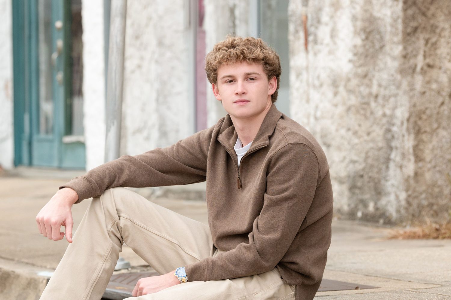 Young man with curly hair sitting outdoors in a brown quarter-zip sweater and khaki pants for a casual portrait.