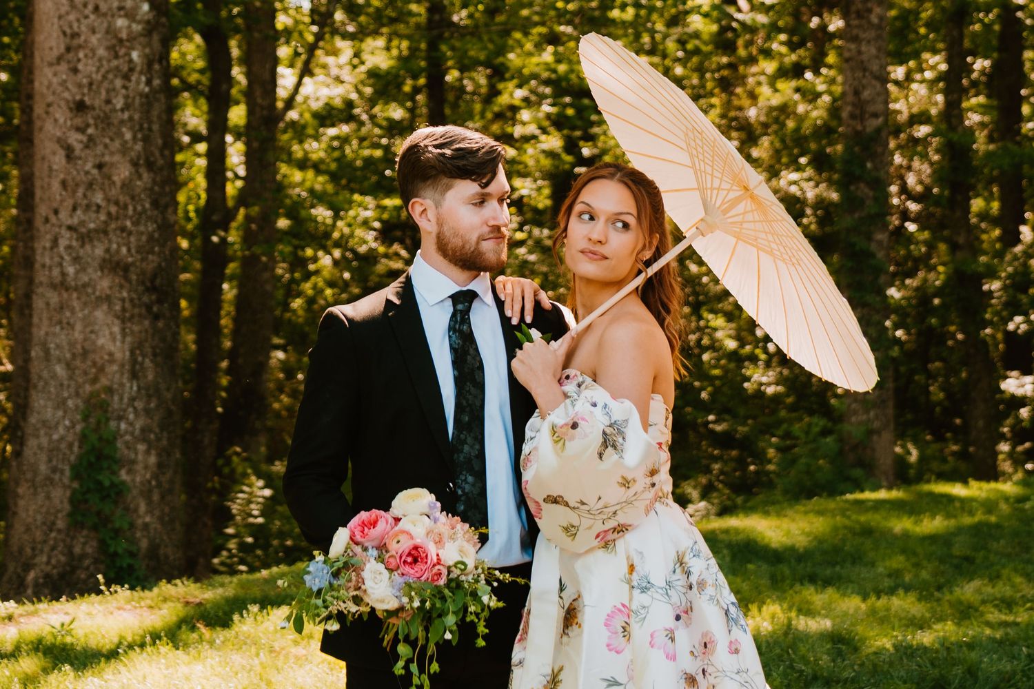 Bride in floral strapless gown holding pink bouquet and parasol stands with groom in black suit outdoors.