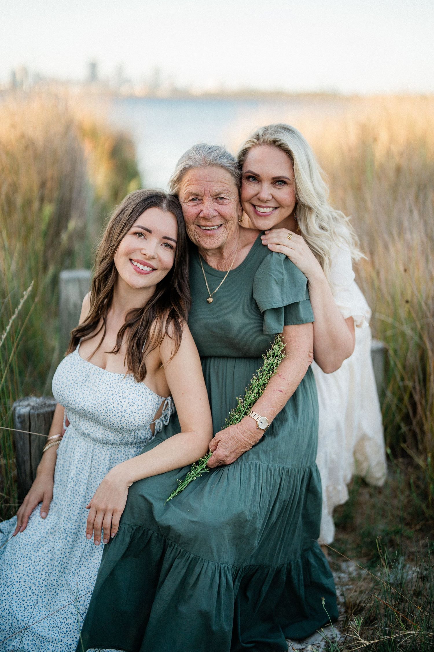 Three generations of women smiling together outdoors near tall grass and water at golden hour.