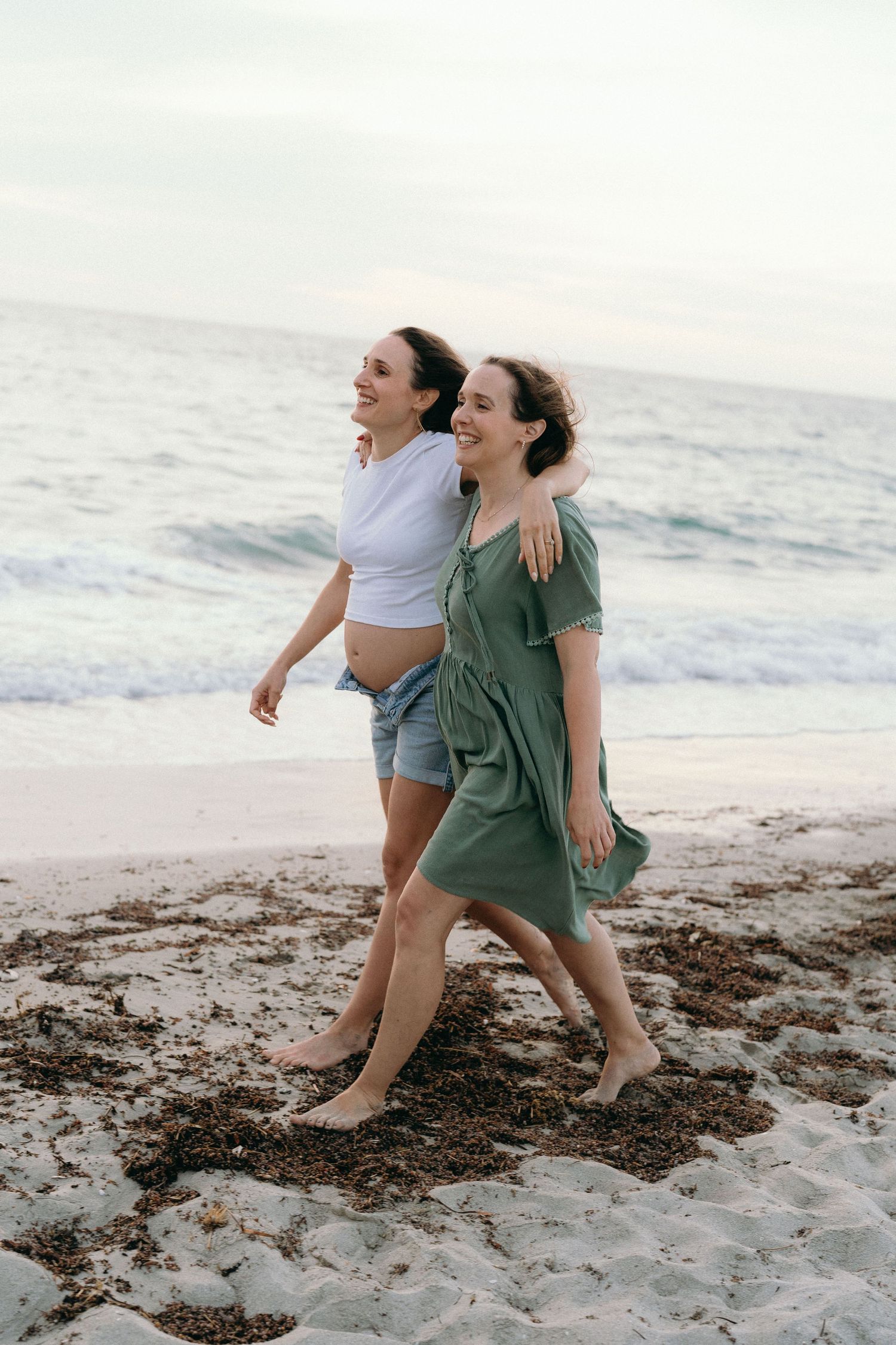 Two women walking barefoot on a sandy beach, one pregnant, smiling and embracing near ocean waves.