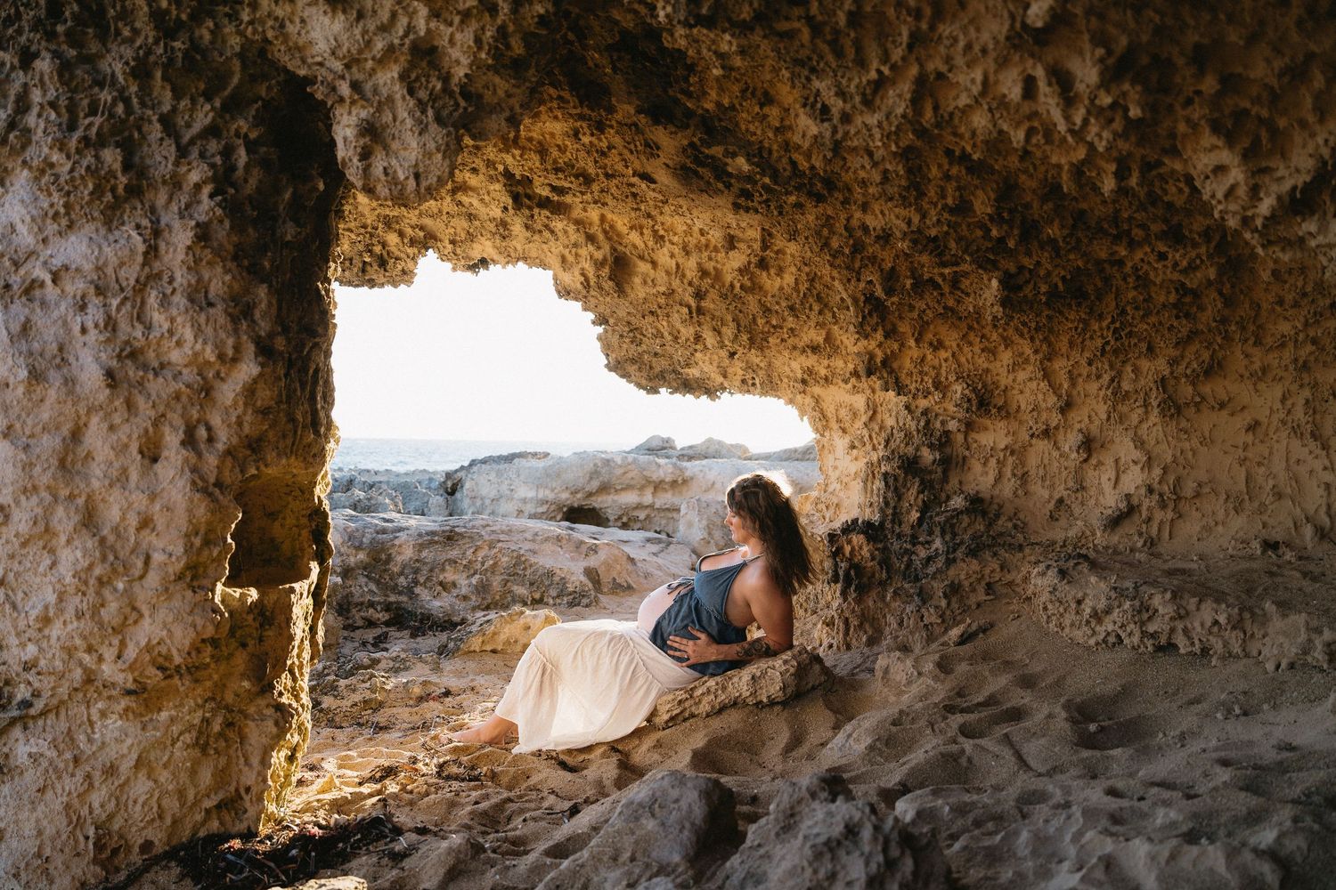 Woman in white skirt sitting inside a rocky cave opening overlooking a sunlit coastal landscape.