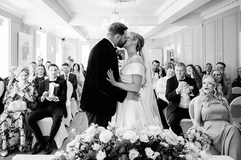 Black and white photo of a bride and groom sharing their first kiss at the altar surrounded by applauding wedding guests.