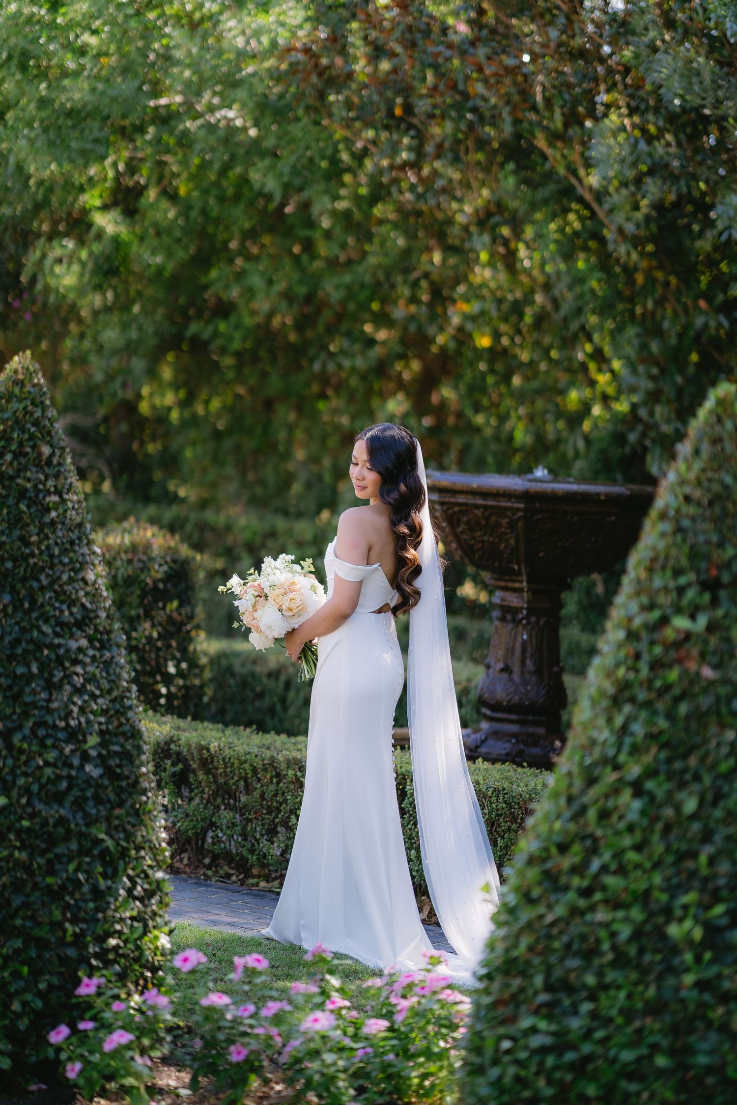 Bride in off-shoulder white gown holding ivory bouquet stands in lush formal garden with manicured hedges and fountain.