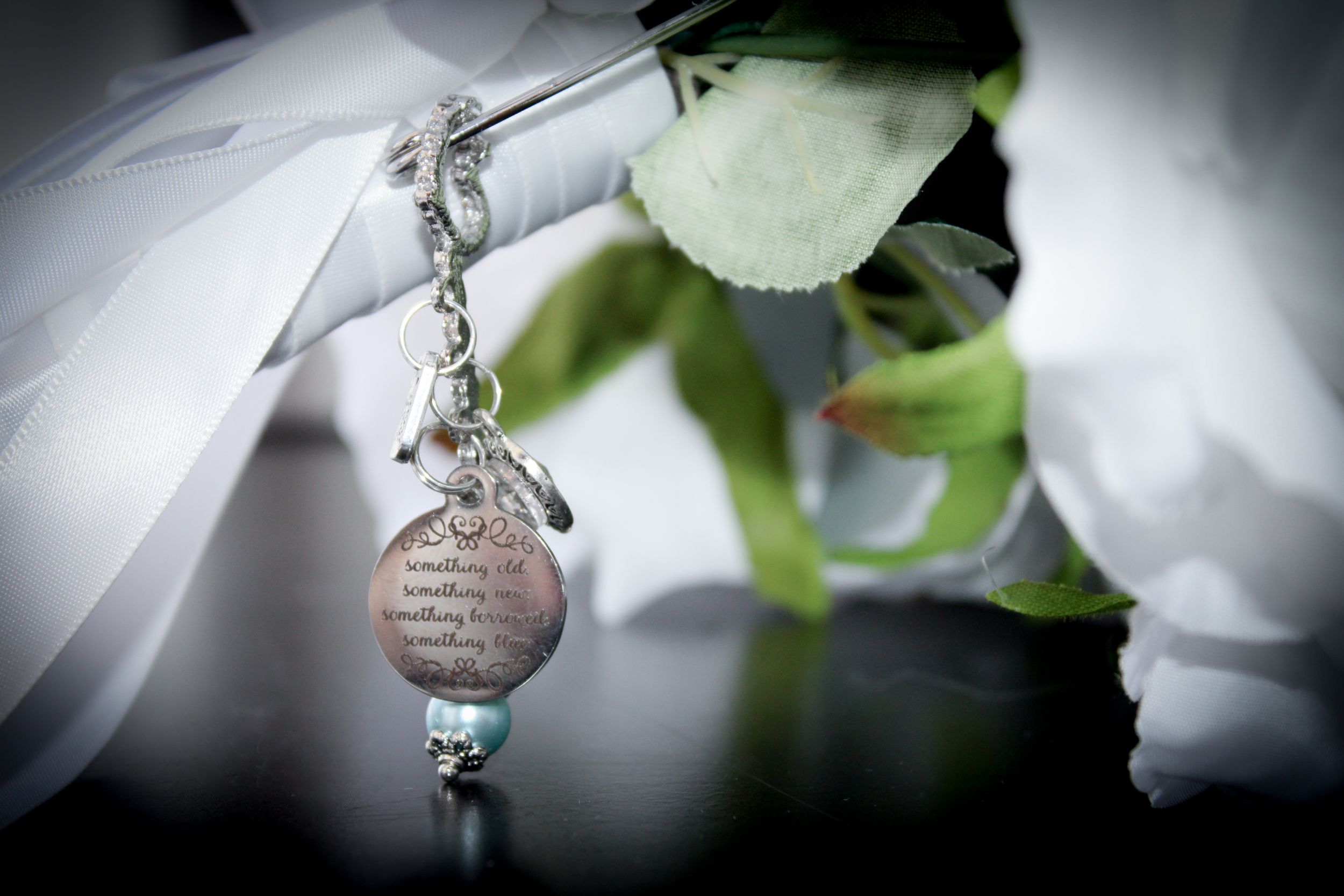 Close-up of a wedding bouquet charm pendant with engraved text and aqua bead, nestled among white roses.