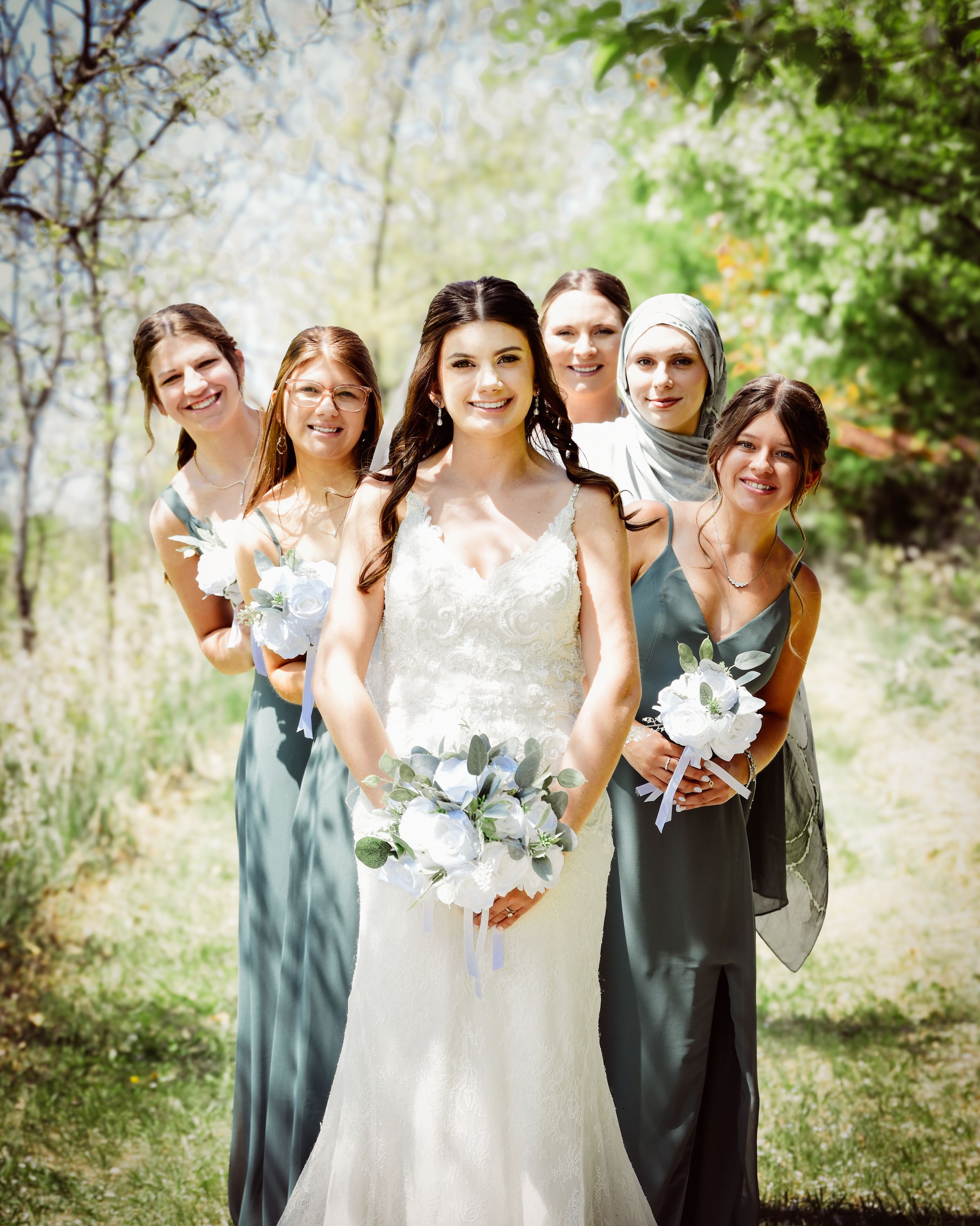 Bride in white lace gown holds bouquet surrounded by bridesmaids in sage green dresses outdoors.