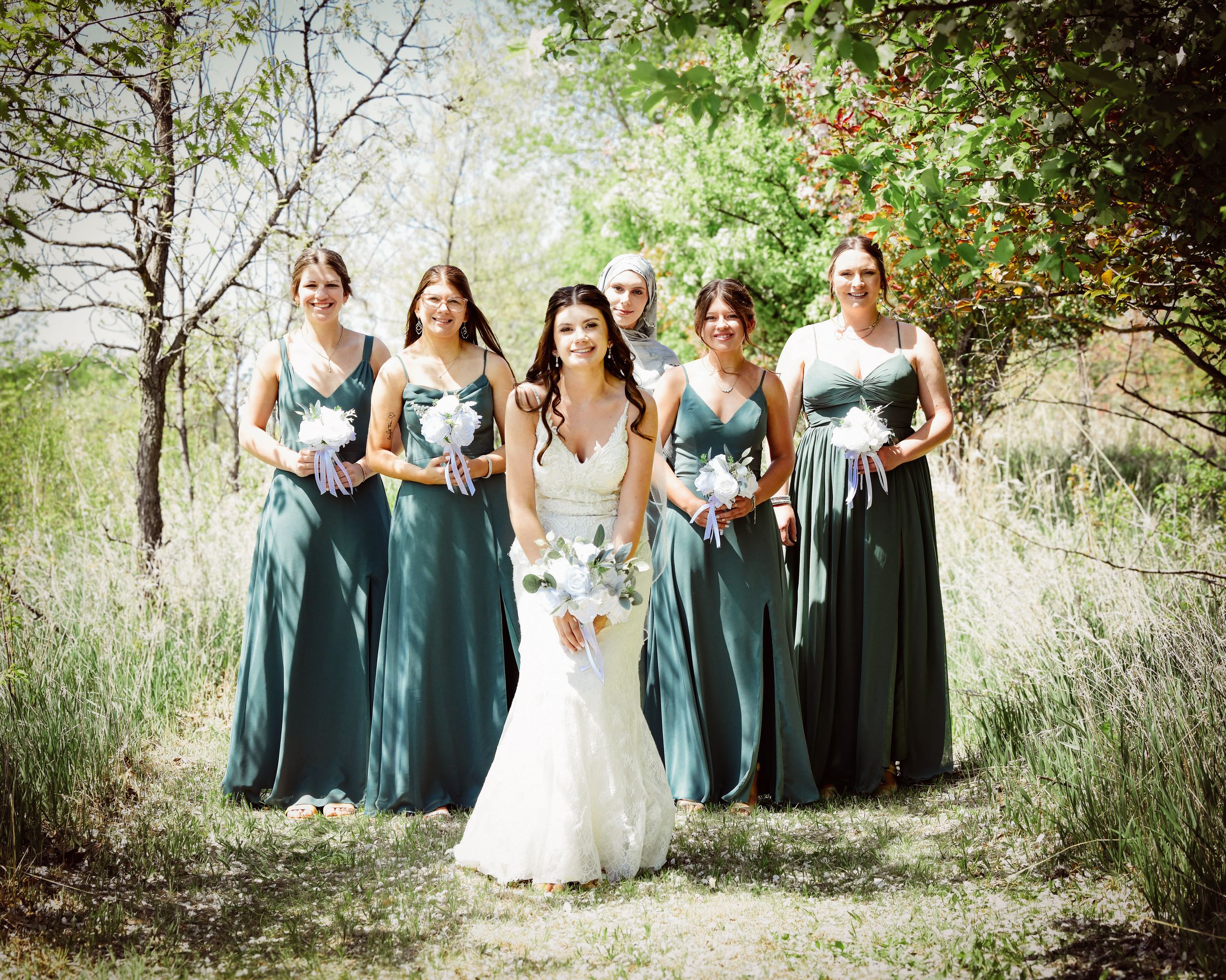 Bride in white gown poses with five bridesmaids in elegant green dresses, holding bouquets outdoors.
