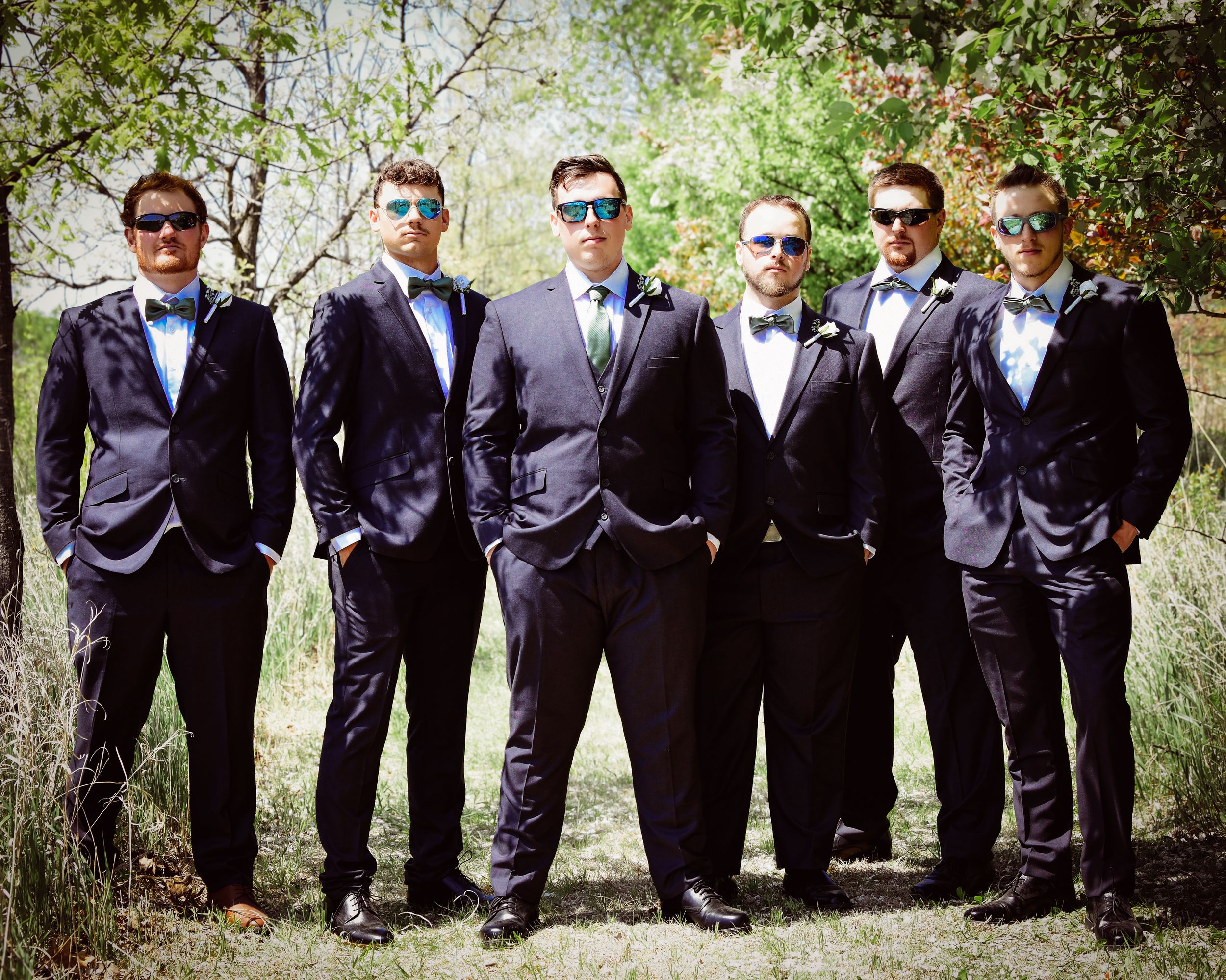 Six groomsmen in matching black tuxedos and blue bow ties wearing sunglasses pose outdoors for a wedding photo.