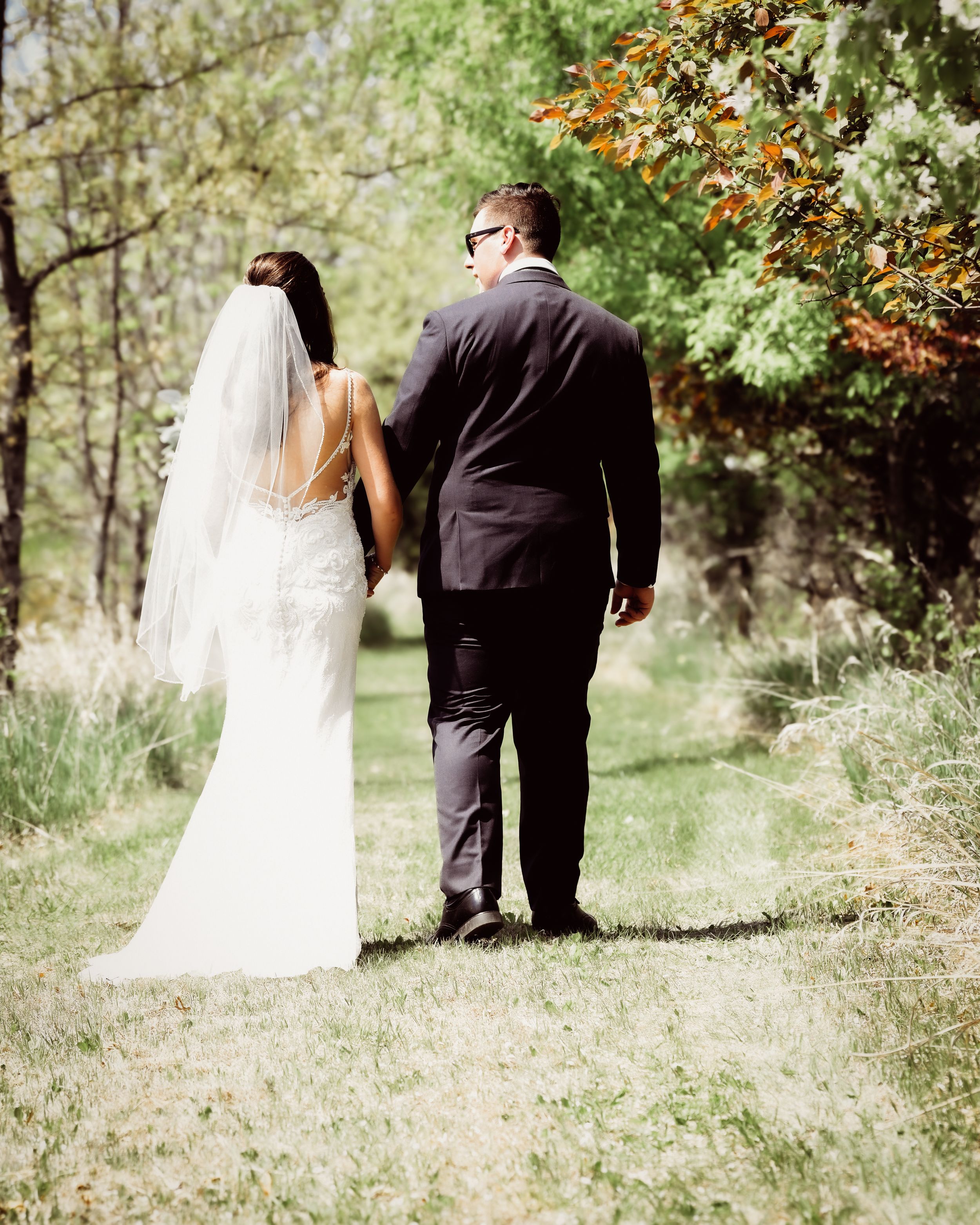 A bride in a white gown and veil walks with a groom in a dark suit through a lush green outdoor setting.