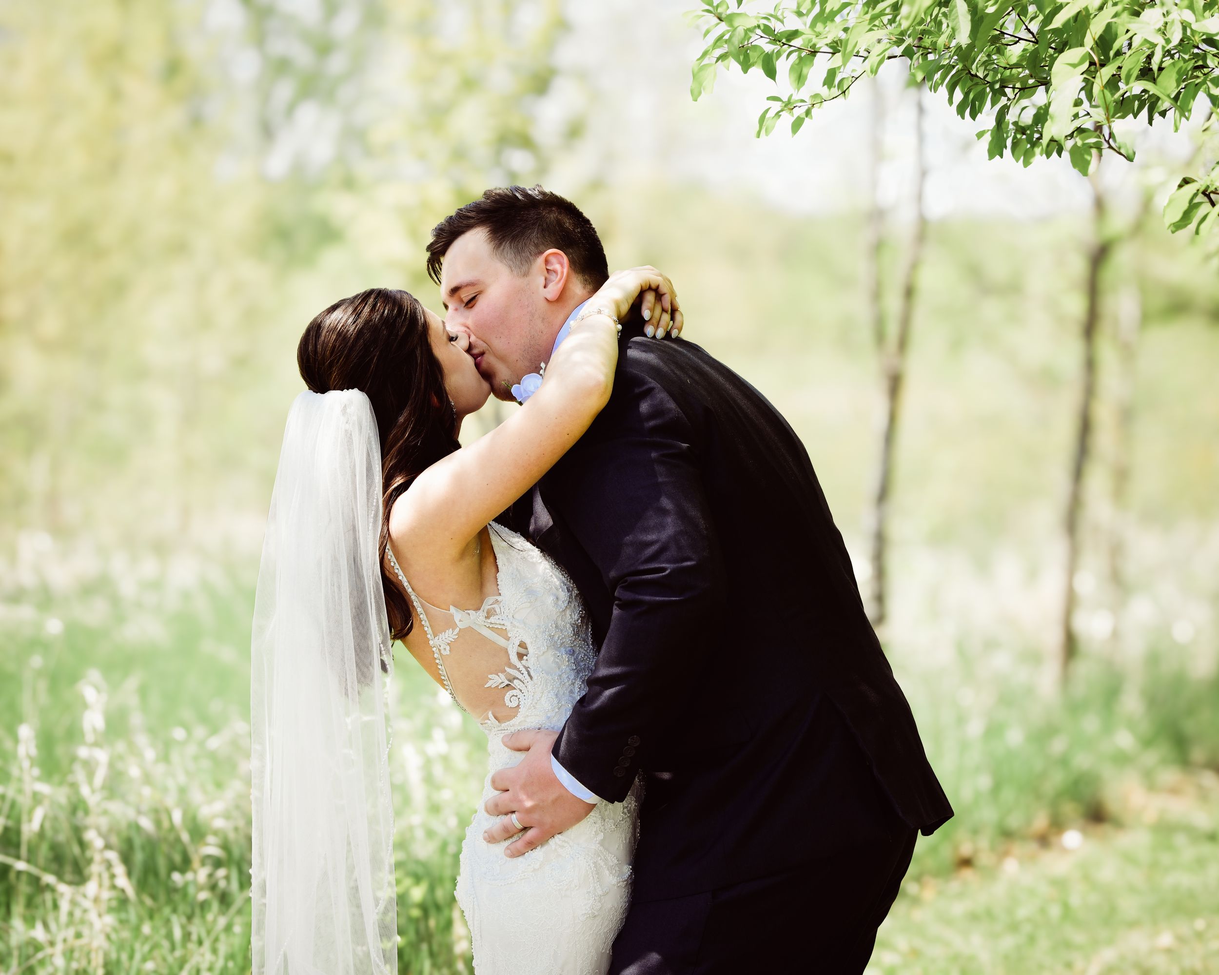 Bride and groom share a romantic kiss outdoors, surrounded by lush green trees on their wedding day.