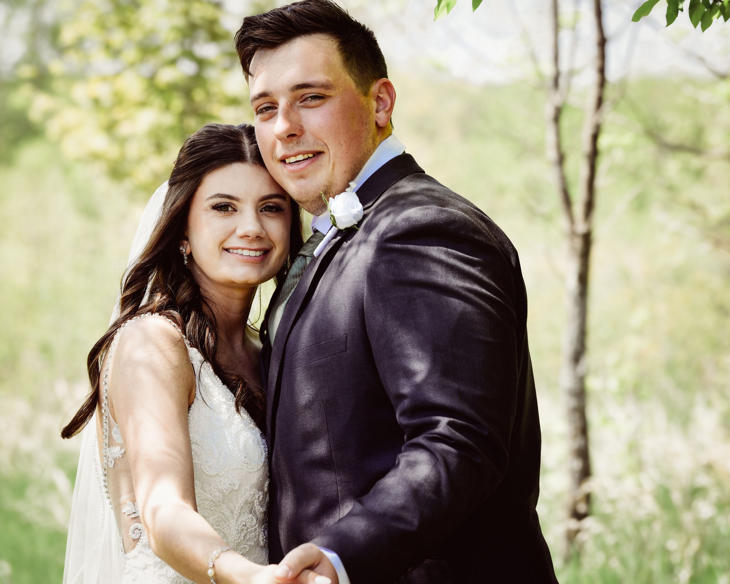 Happy couple in wedding attire embracing outdoors, bride in white lace gown and groom in dark suit, surrounded by greenery.