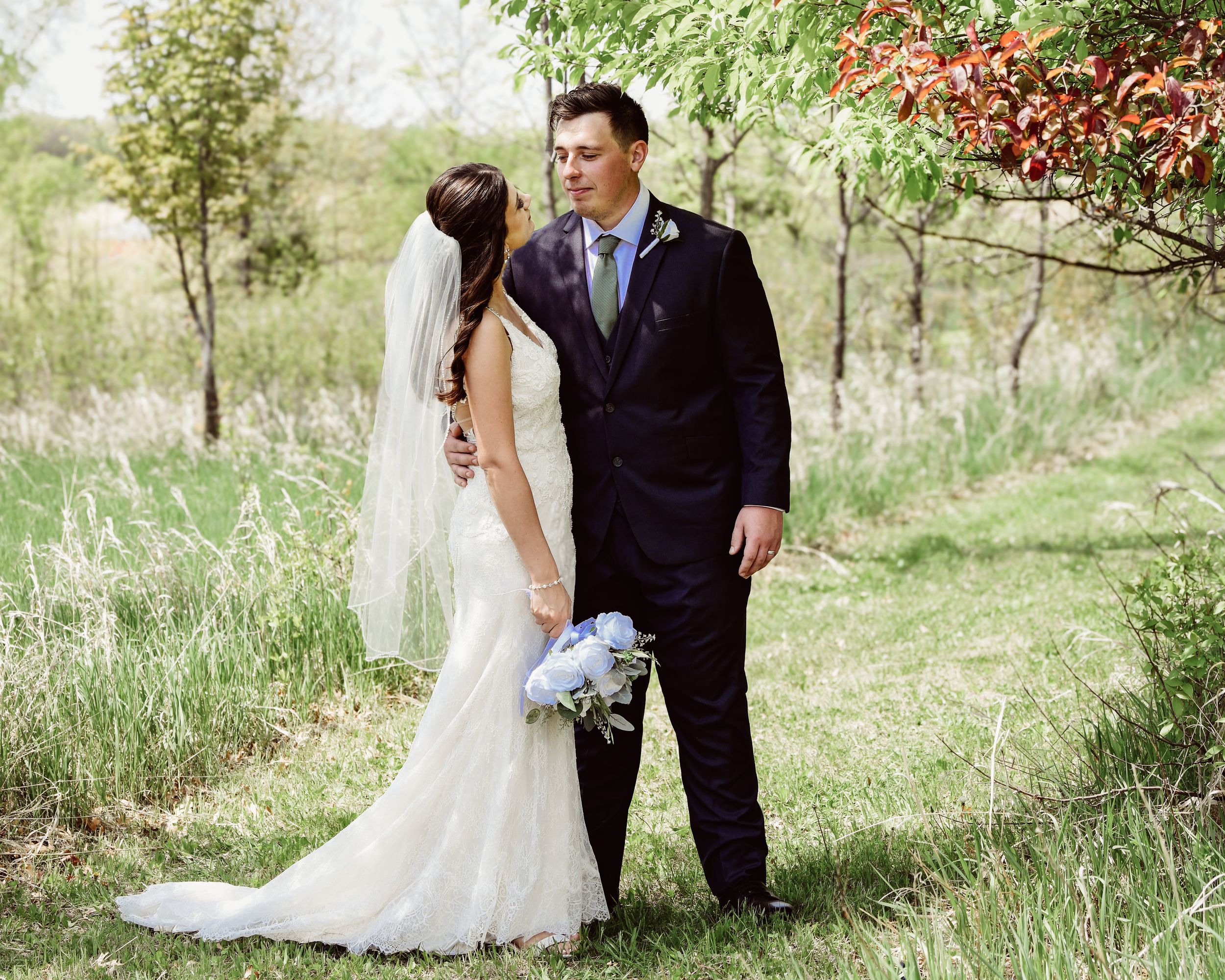 Bride and groom share a romantic moment outdoors, surrounded by lush greenery, as she holds a white bouquet in a elegant wedding gown.