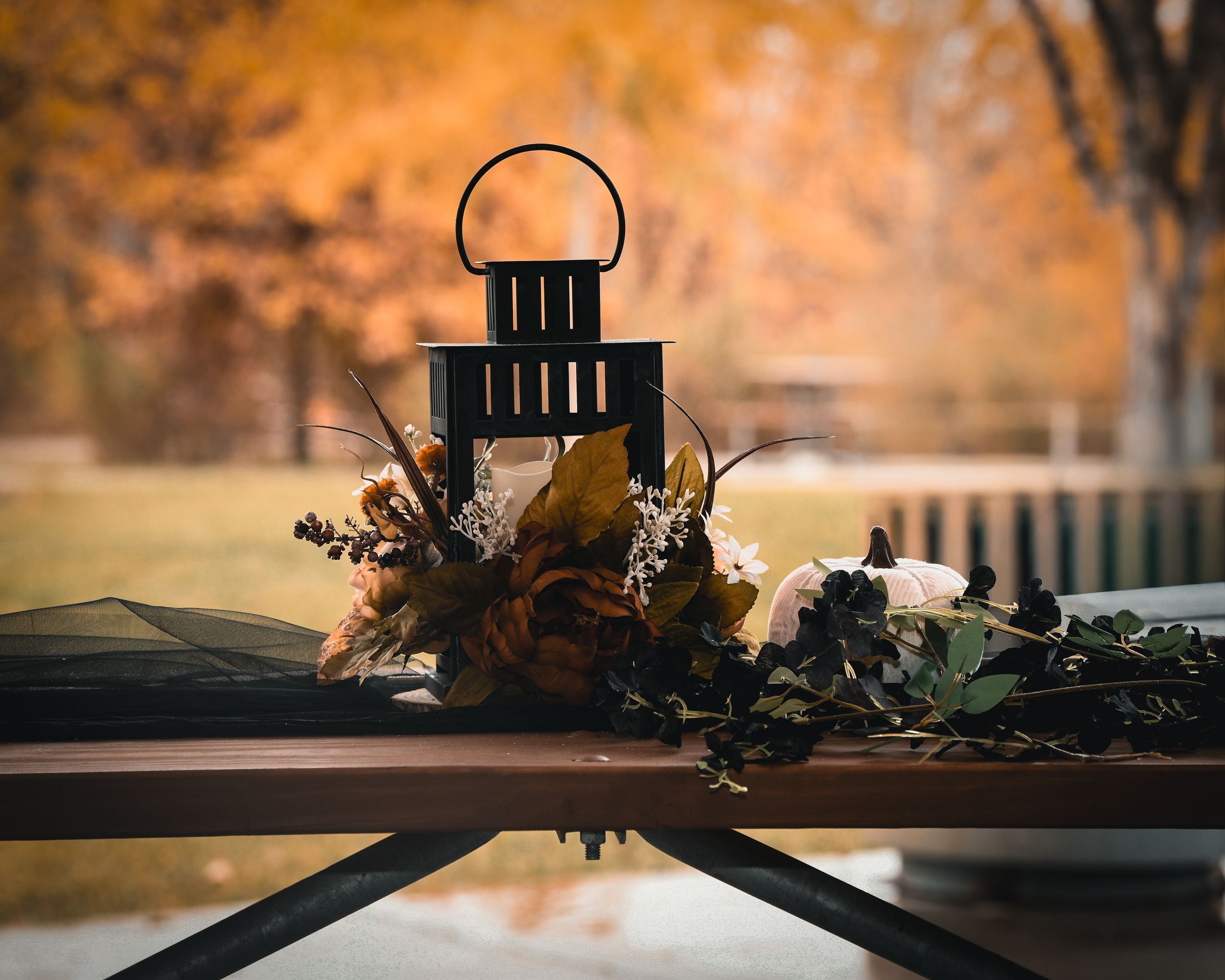 Black lantern centerpiece with autumn floral arrangement on table, fall foliage in background.