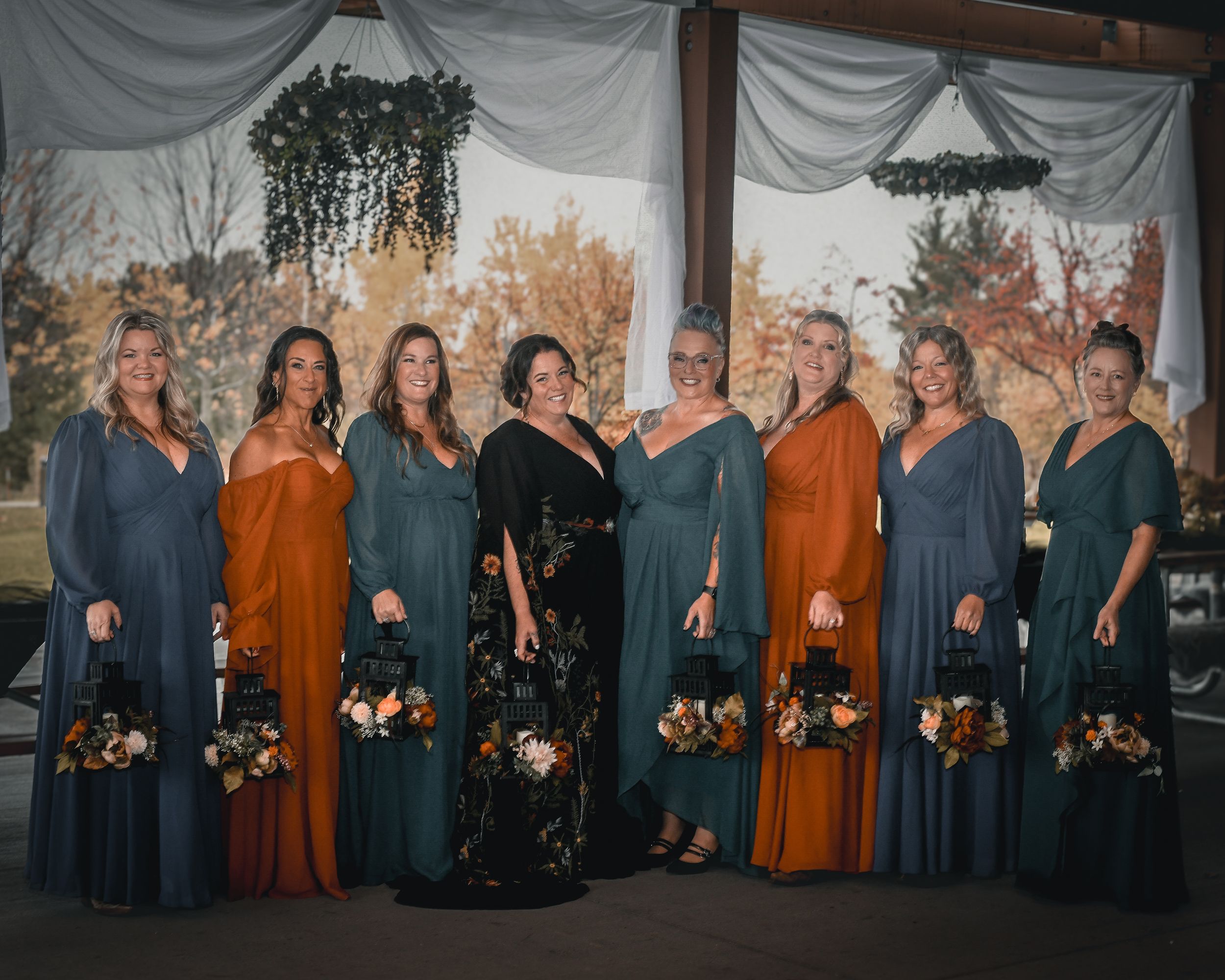 Wedding party posing in teal and burnt orange bridesmaid dresses with floral bouquets at an outdoor fall wedding.