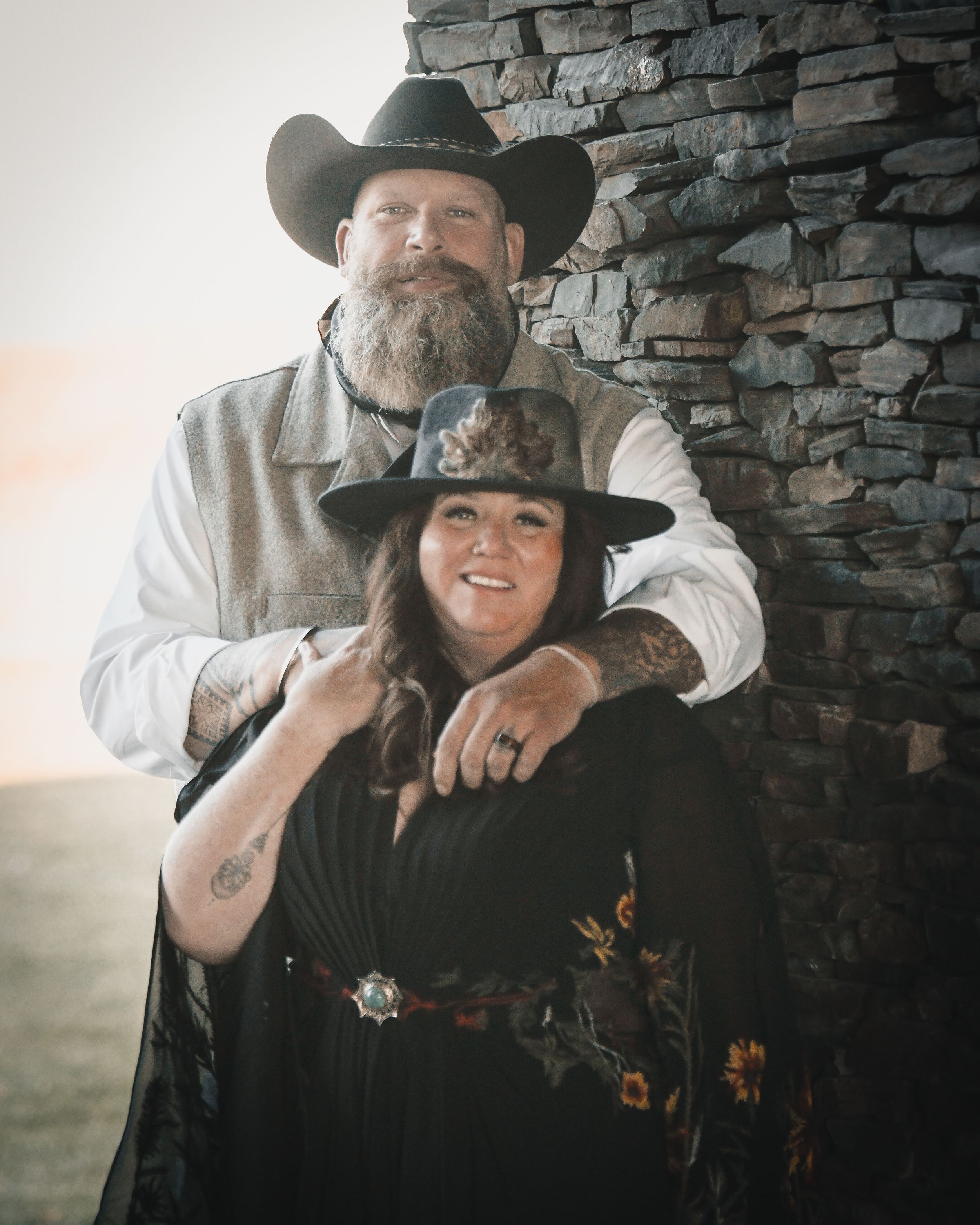 Couple in western cowboy hats and rustic attire pose together against a stone wall.