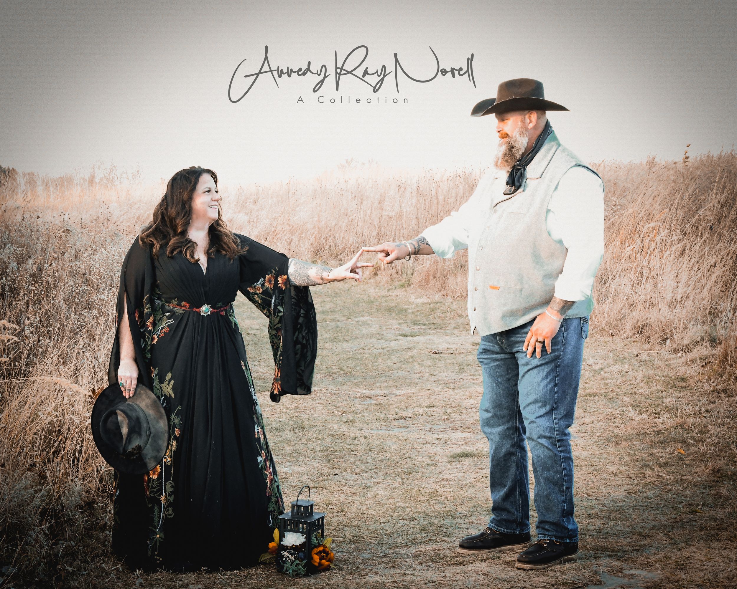 A woman in a black floral gown and a man in a cowboy hat stand in a golden field during a rustic outdoor photoshoot.
