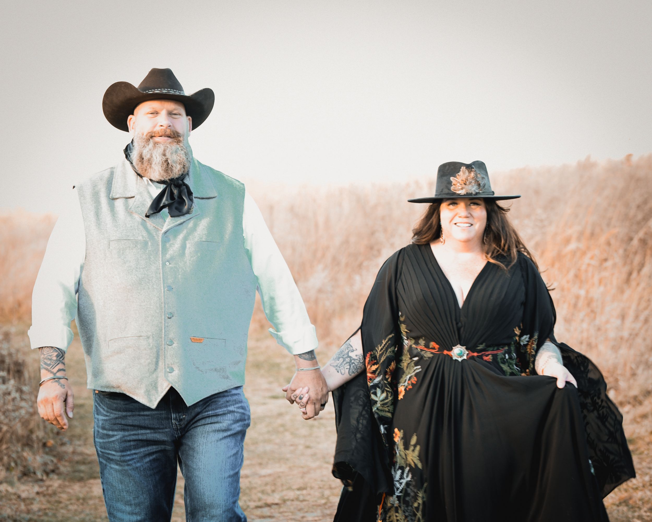 Couple in western-style attire holding hands while walking through a golden field in cowboy hats.