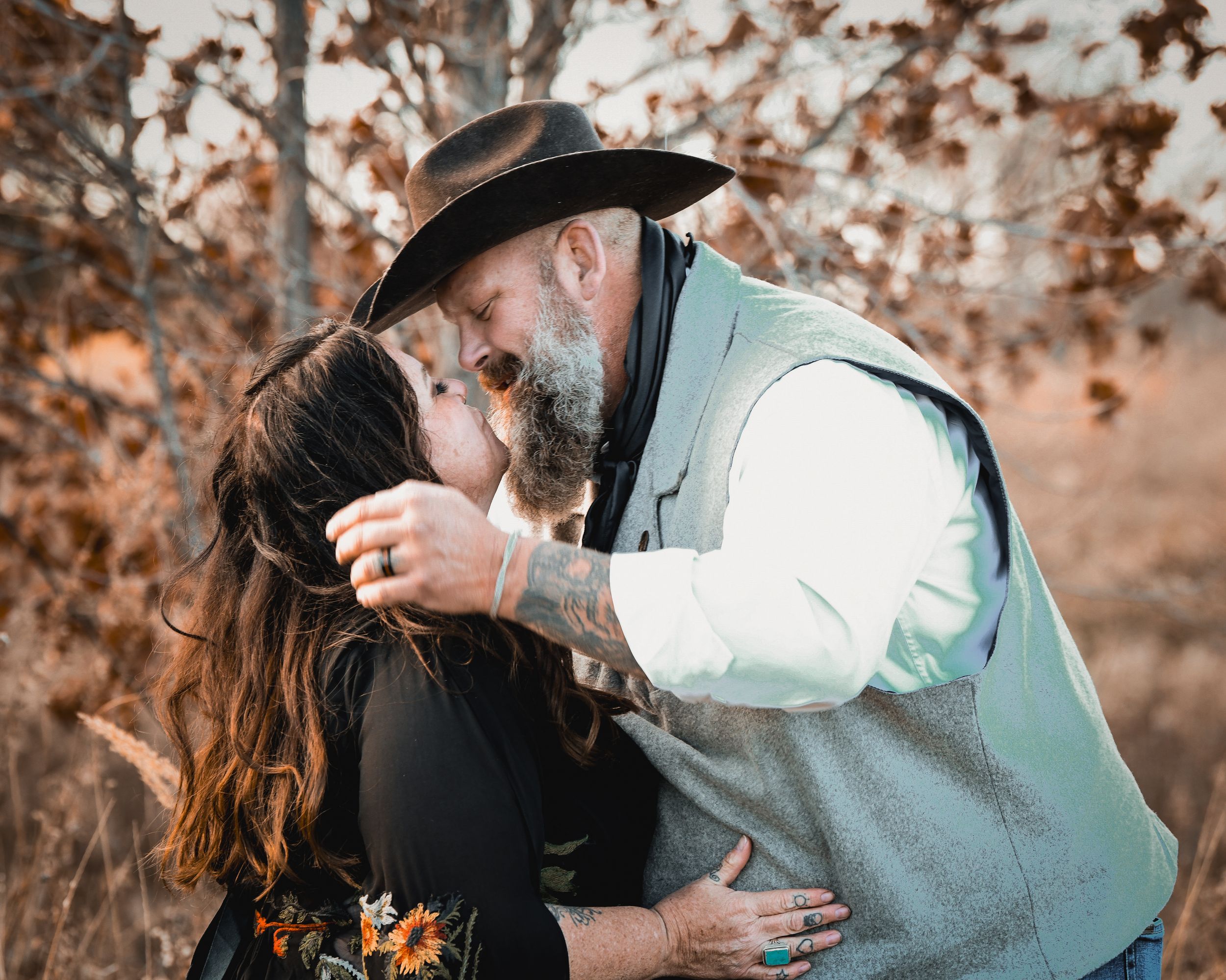 A bearded man in a cowboy hat and vest shares a romantic kiss with a woman outdoors in autumn.