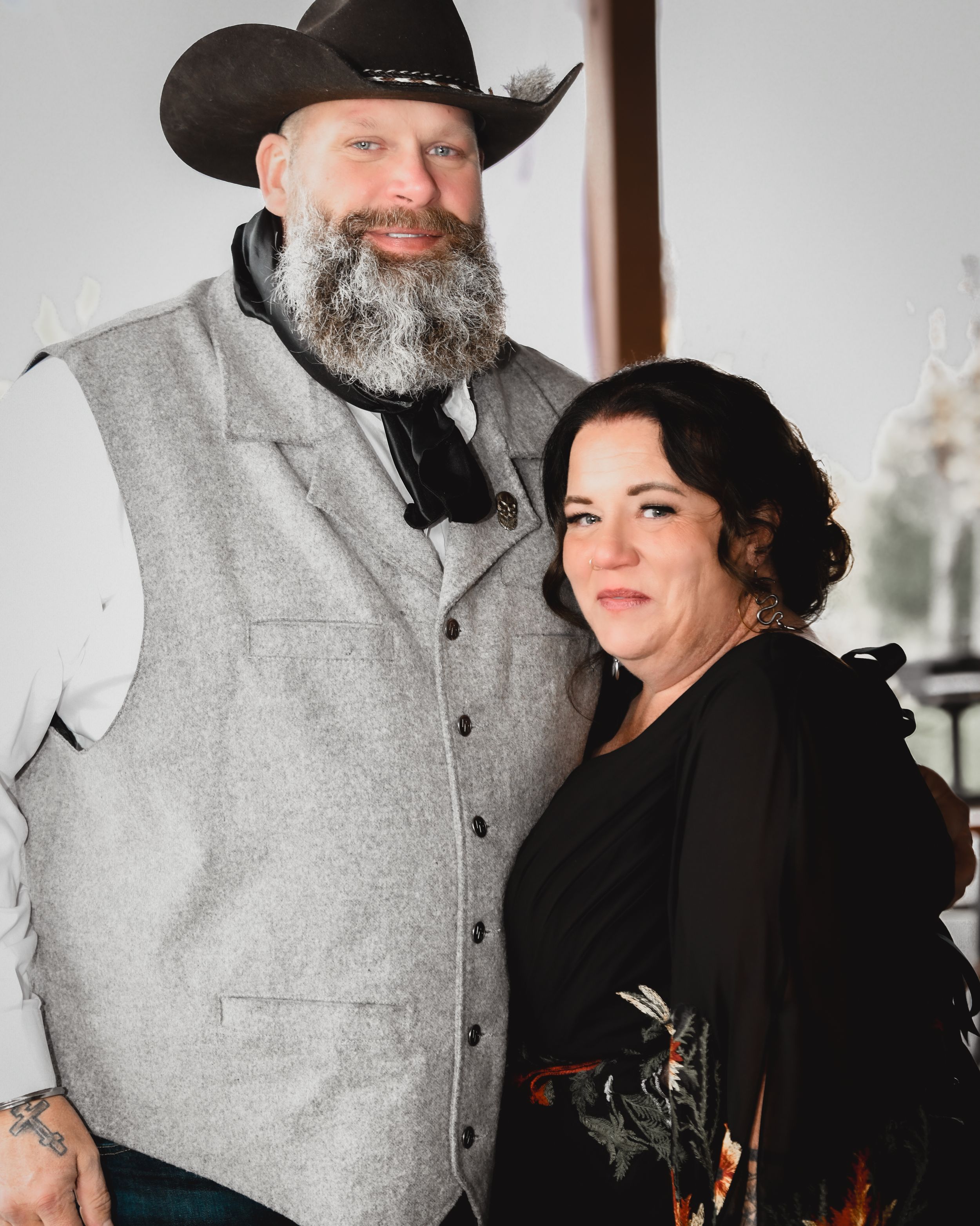 Smiling couple posing together at a wedding, man wearing a gray vest and cowboy hat with a bearded smile.