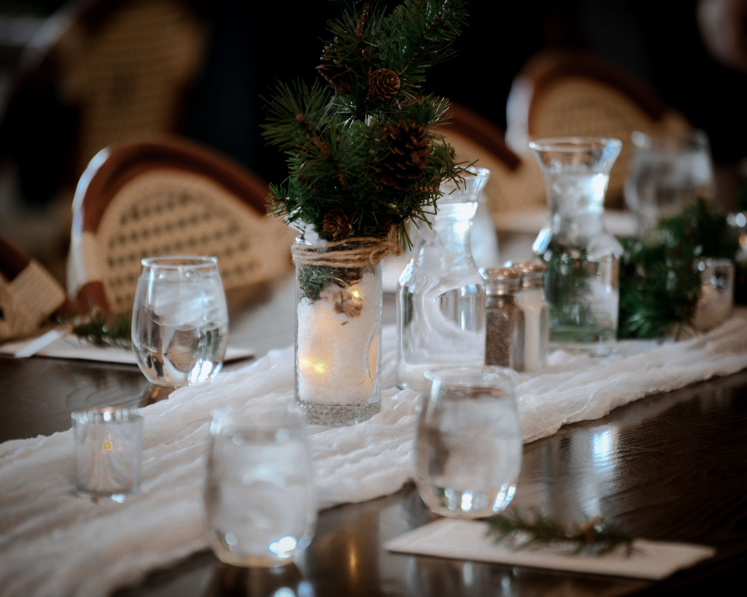 Elegant winter wedding table centerpiece with candles, pine greenery, and glassware on a white linen runner.