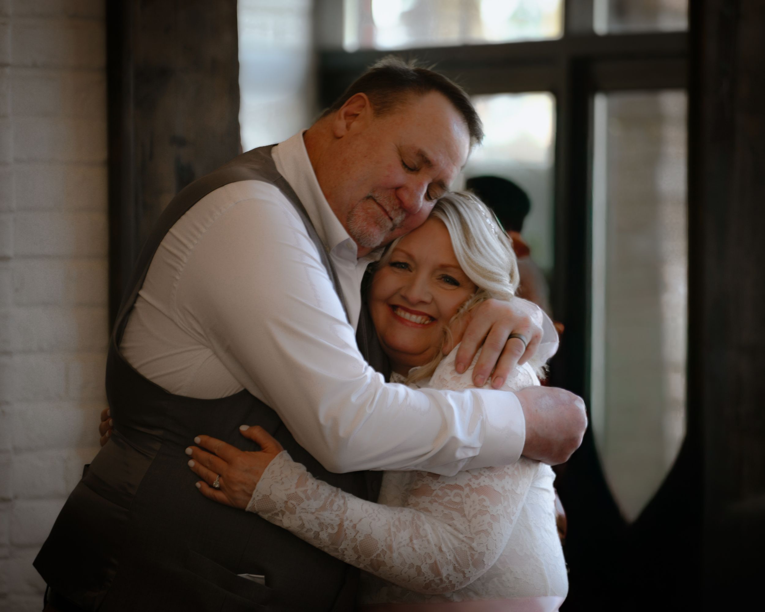 A smiling bride in white lace embraces a man in a gray vest during a warm wedding celebration.