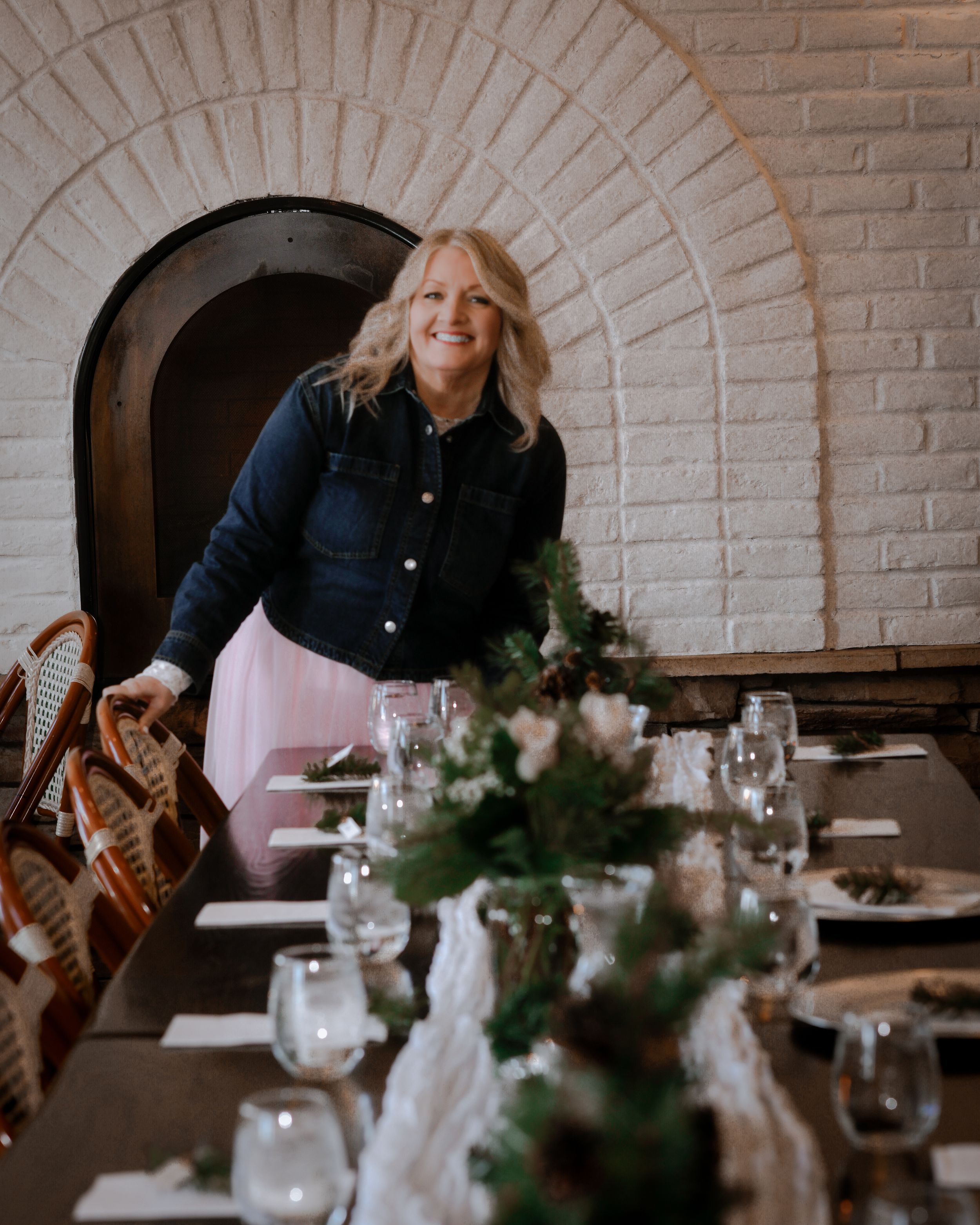 Smiling blonde woman in denim jacket and pink skirt arranging a decorated dining table in a brick-arched venue.