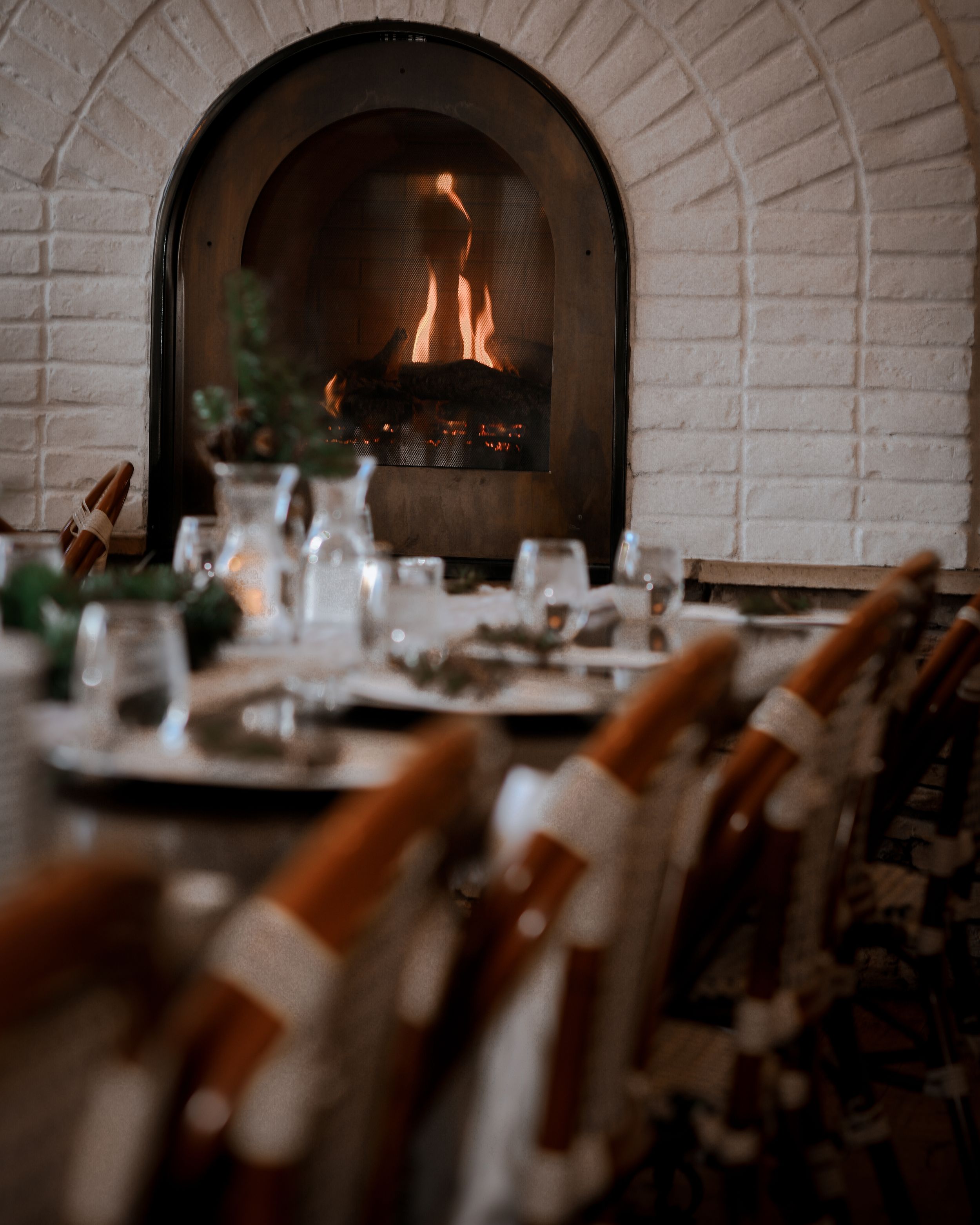 Elegant wedding reception table set with glassware and white decor beside a glowing arched brick fireplace.