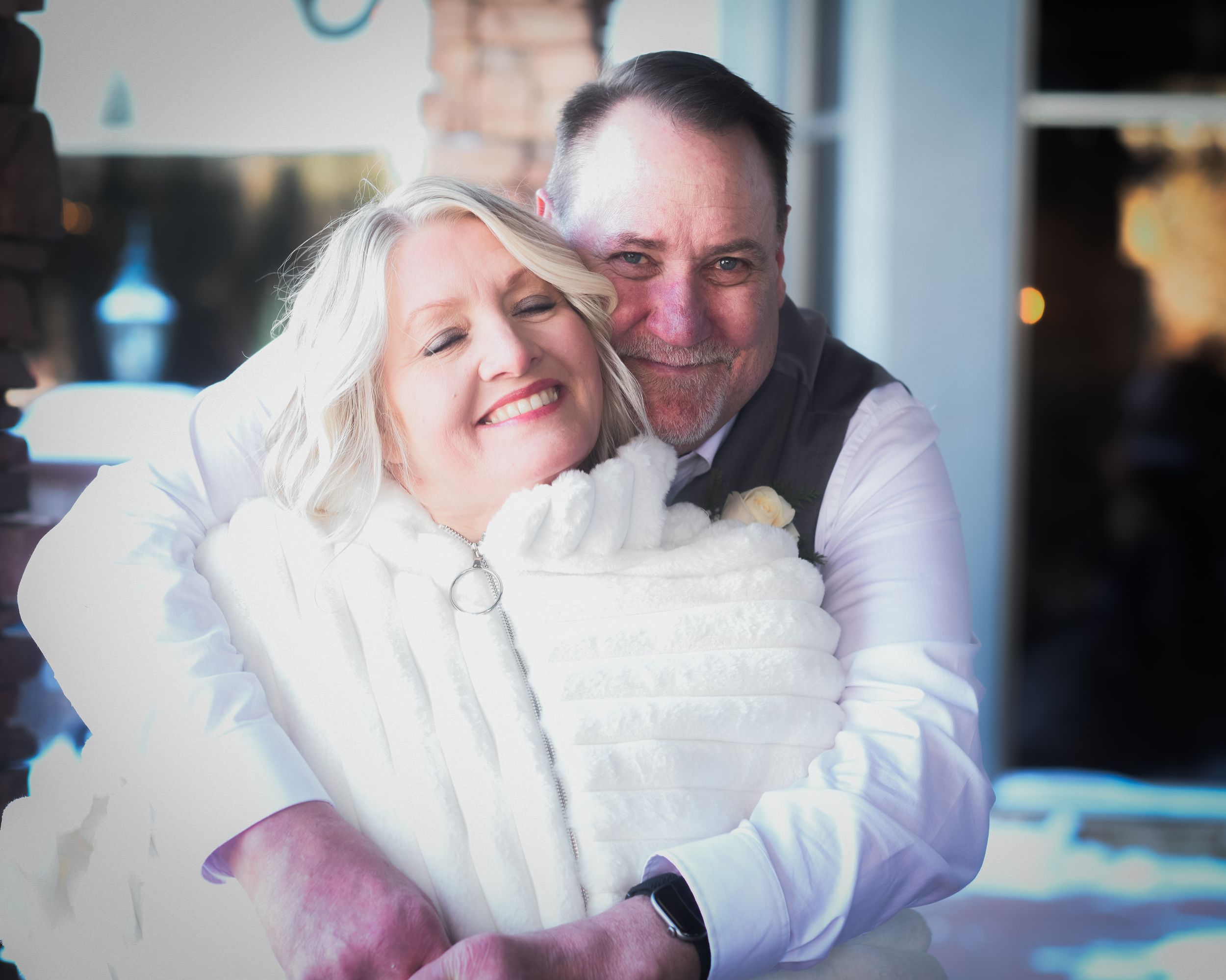 A joyful couple embraces outdoors, the woman in a white fur coat and the man in a gray vest.