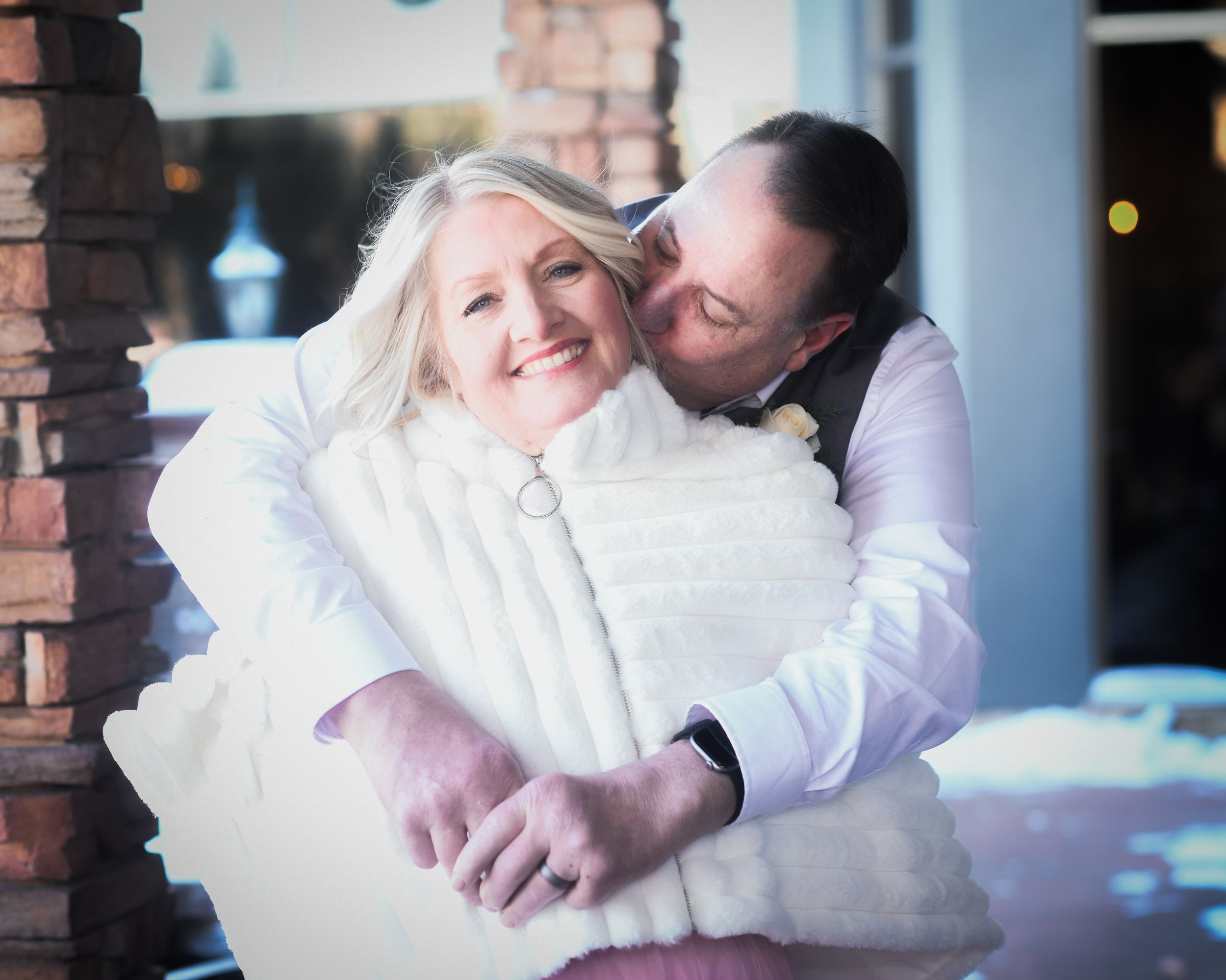 A groom embraces and kisses his smiling blonde bride wearing a white fur wrap at a winter wedding celebration.