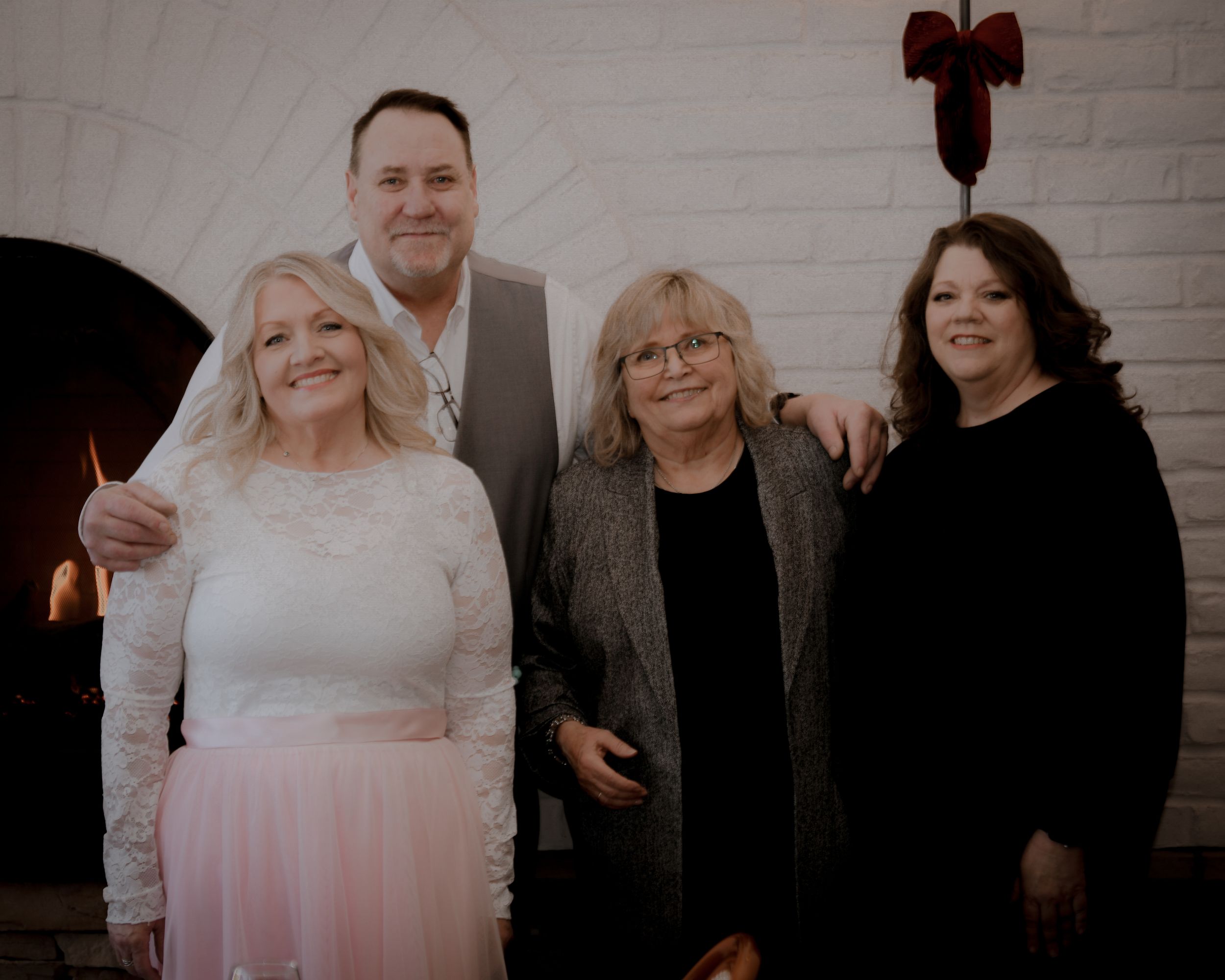 Group of four adults posing together near a fireplace, with a woman in a white lace wedding dress and pink skirt.