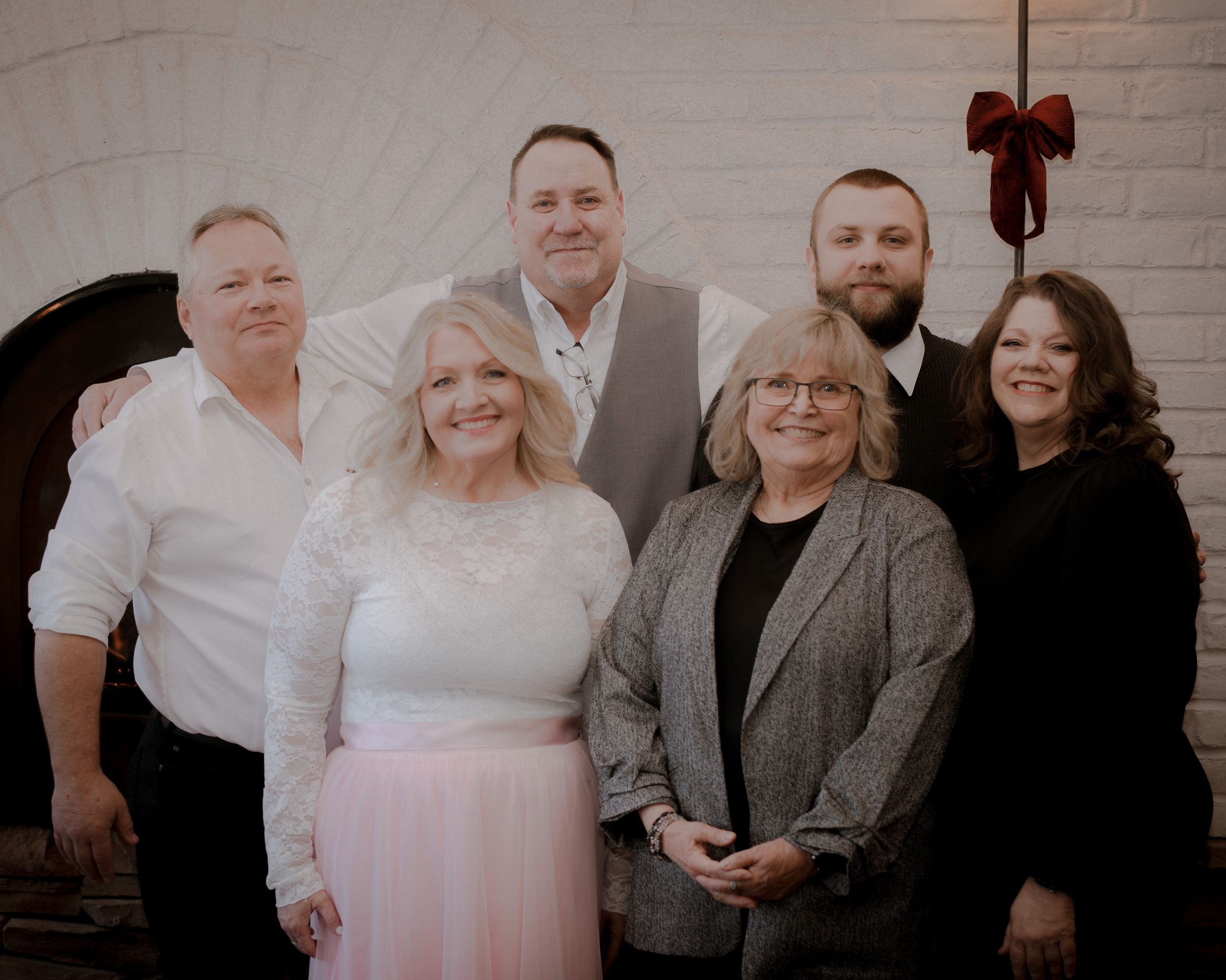 Six smiling family members pose together in formal attire at an elegant white brick venue with holiday decor.