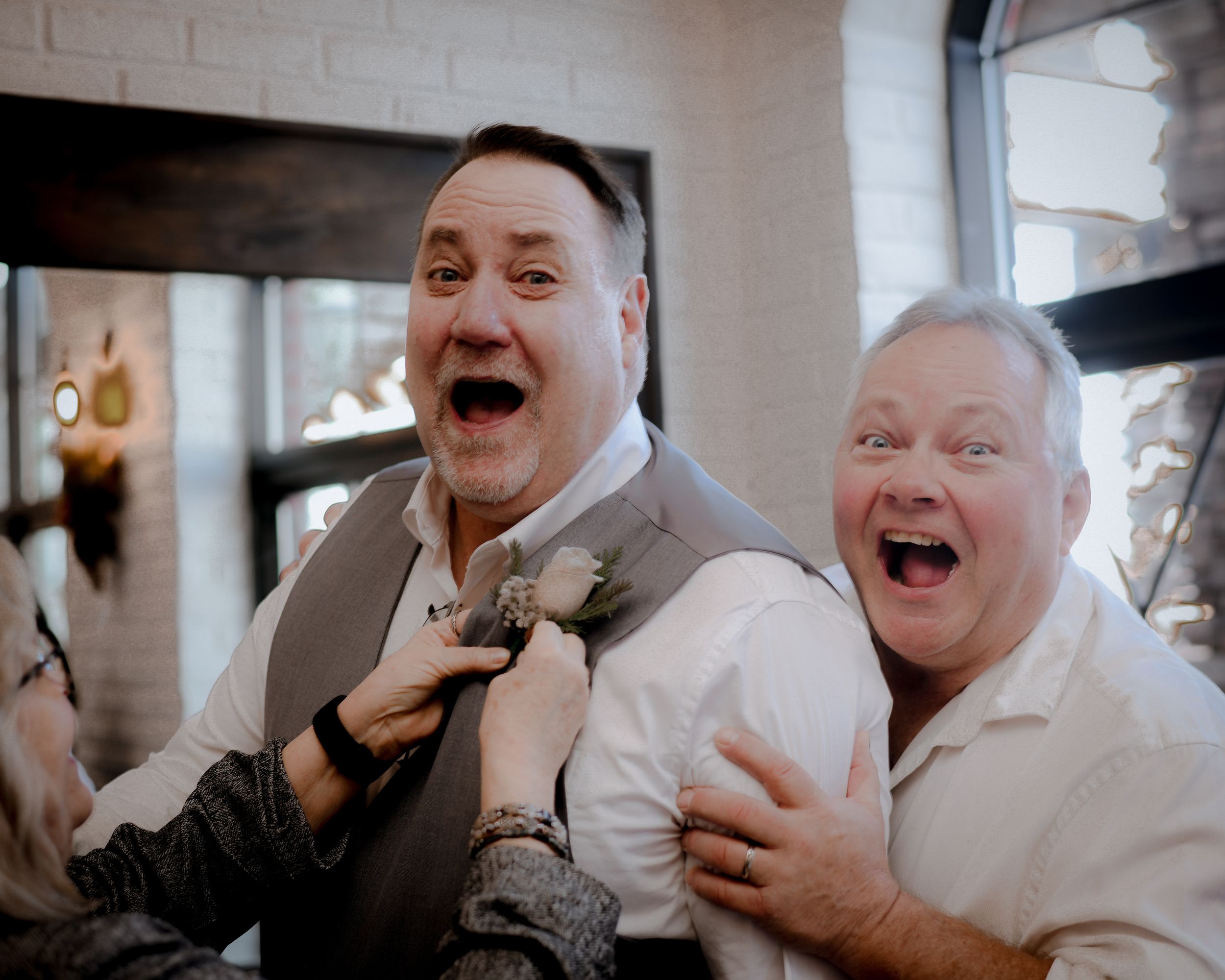Two joyful men laughing and celebrating together at a wedding reception, one wearing a gray vest with a boutonniere.