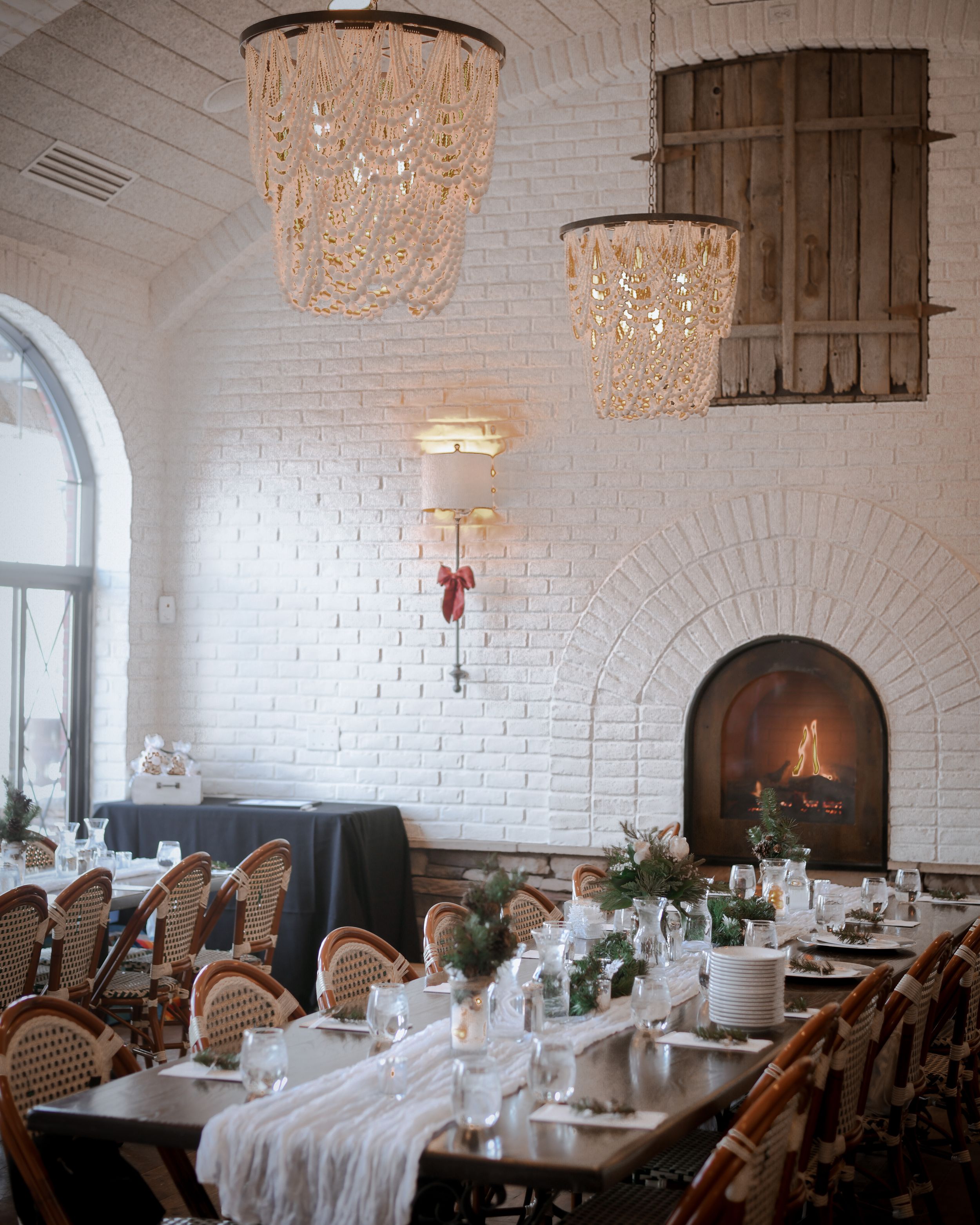 Elegant wedding reception venue with white brick walls, crystal chandeliers, long dining tables, and a glowing fireplace.