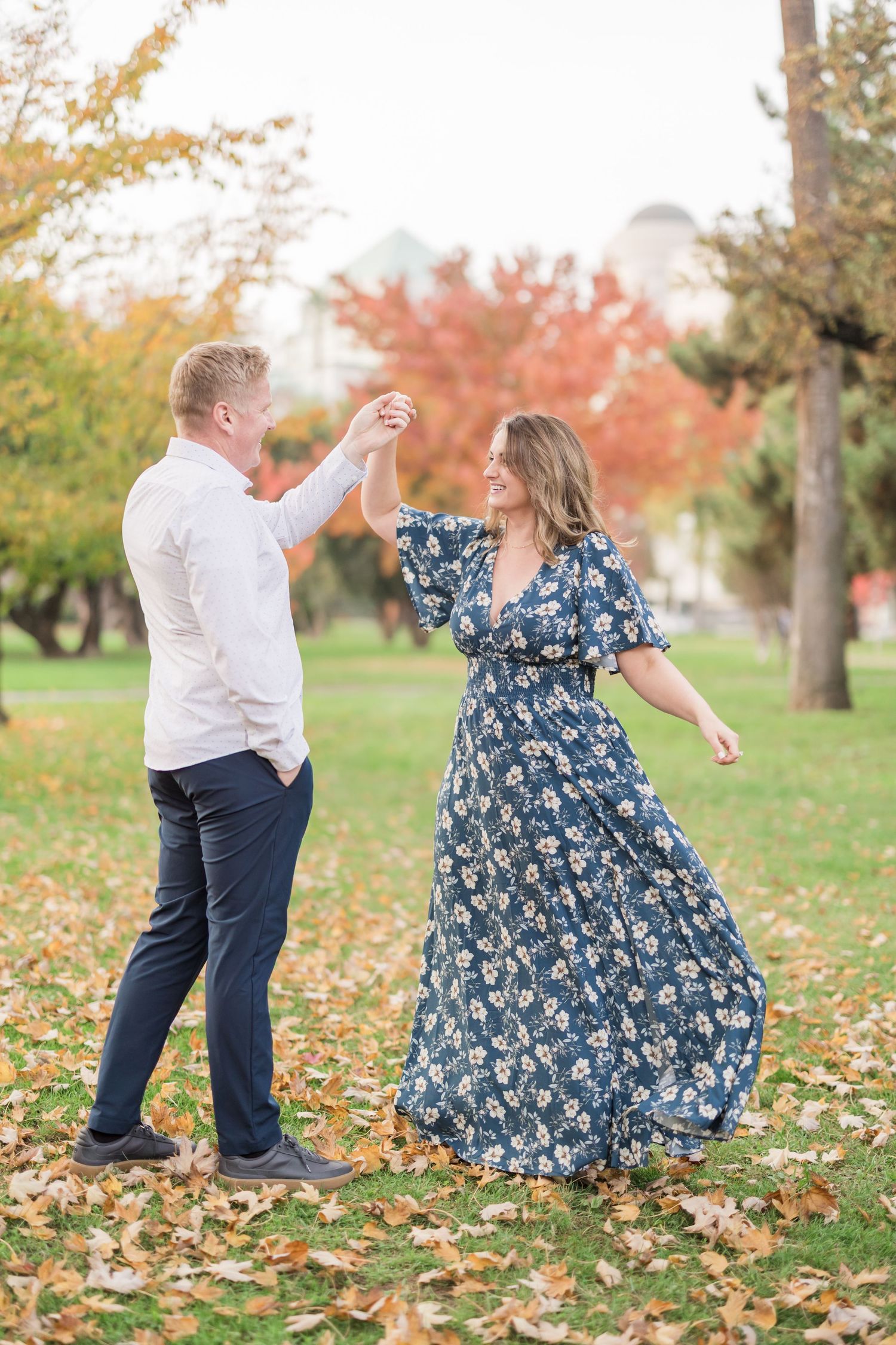 Couple dancing outdoors in autumn park, woman in blue floral maxi dress twirling, man in white shirt and navy pants.