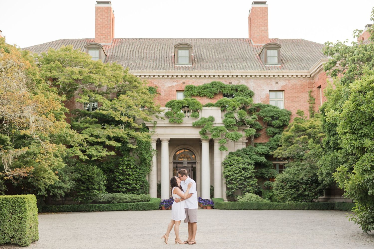 Couple embracing in front of elegant brick mansion with lush greenery and white columns at garden estate.