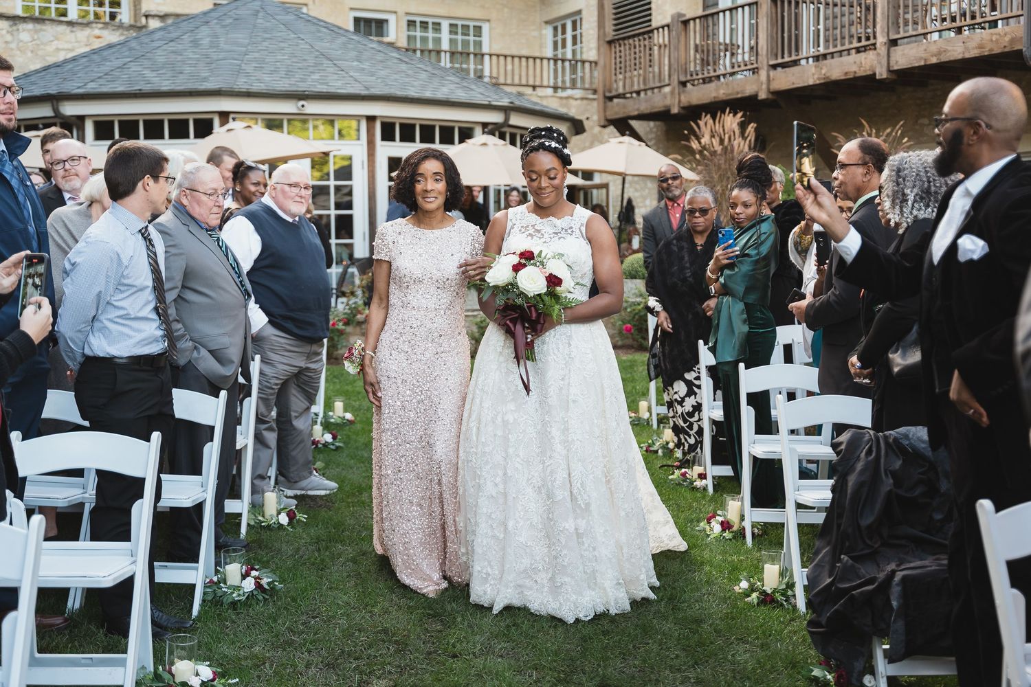 Two brides walking down the aisle at outdoor wedding ceremony, guests applauding, one in gold gown, one in white ball gown.