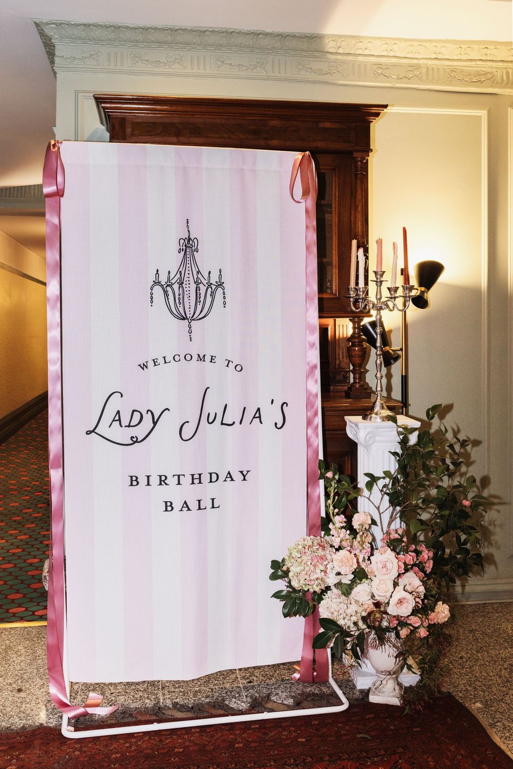 Pink striped welcome banner for Lady Julia's Birthday Ball, adorned with chandelier graphic and pink ribbon ties.