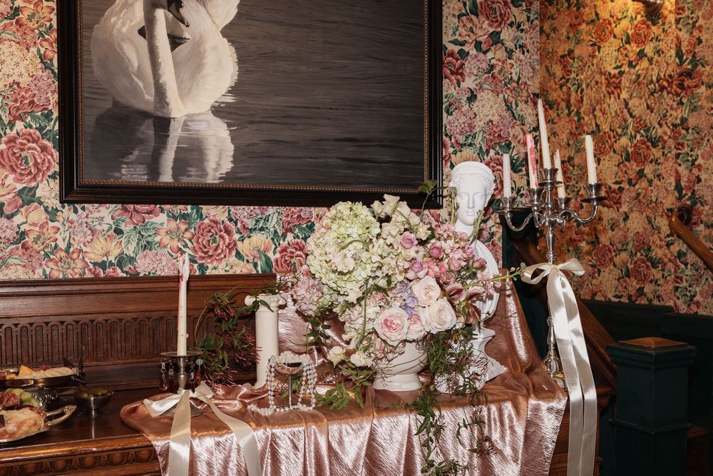 Elegant wedding table with floral arrangement, candelabra, pink roses, and floral wallpaper backdrop in a vintage-styled venue.