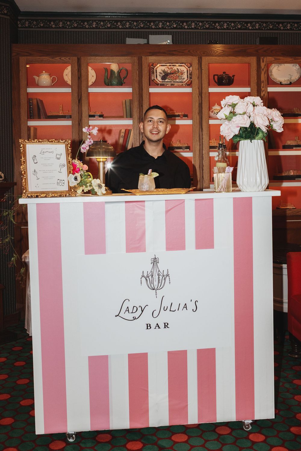 Bartender smiling behind a pink and white striped Lady Julia's Bar counter with floral decorations.