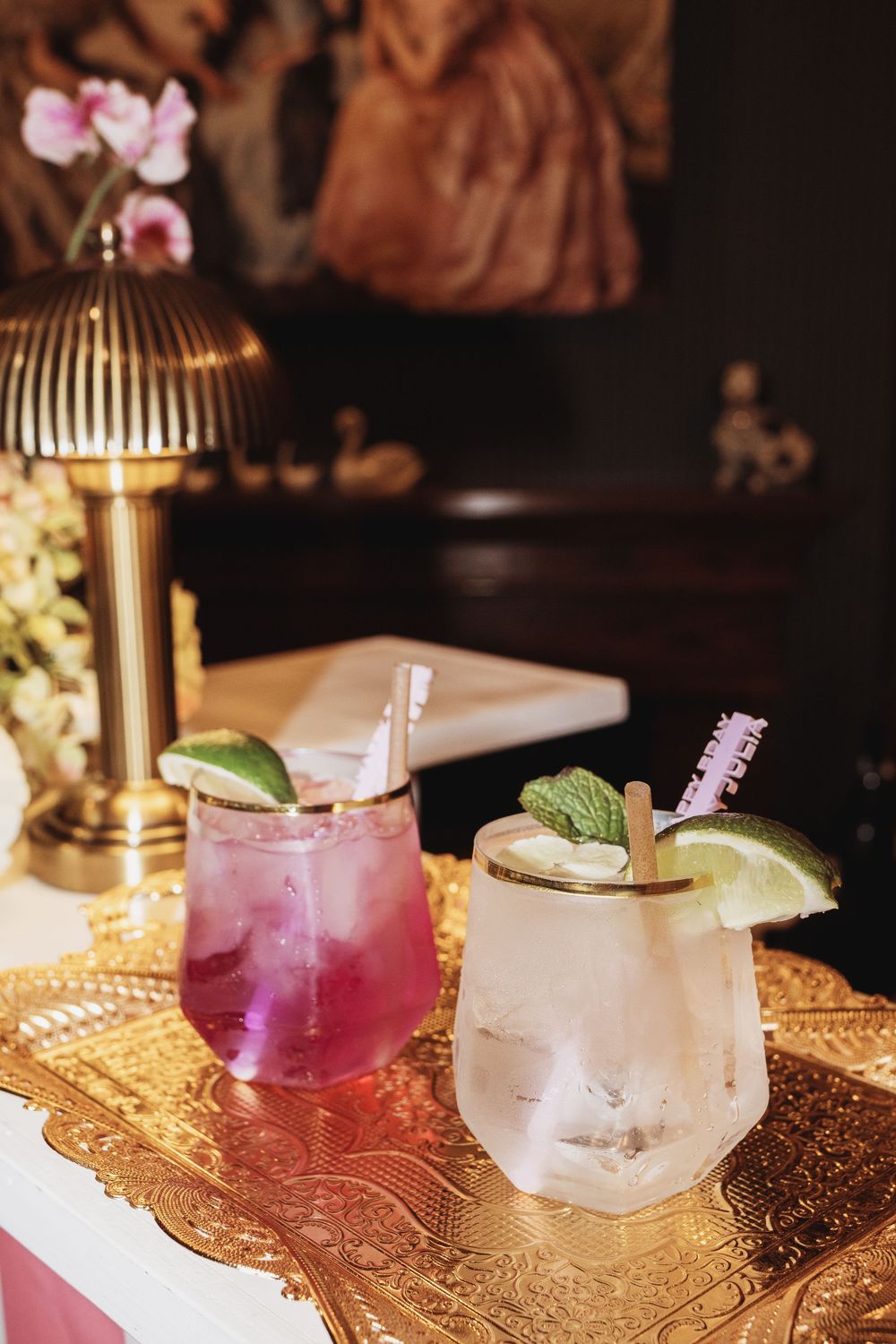 Two elegant cocktails with lime garnishes on a golden tray beside a brass lamp in a moody bar setting.