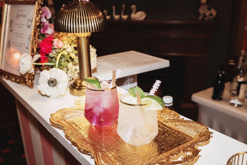 Two colorful cocktails on a gold tray at an elegant bar with floral decor and a brass lamp.