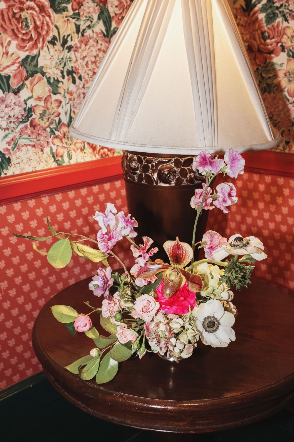 Elegant floral arrangement with pink orchids and anemones on a wooden table beside a decorative lamp with floral wallpaper.