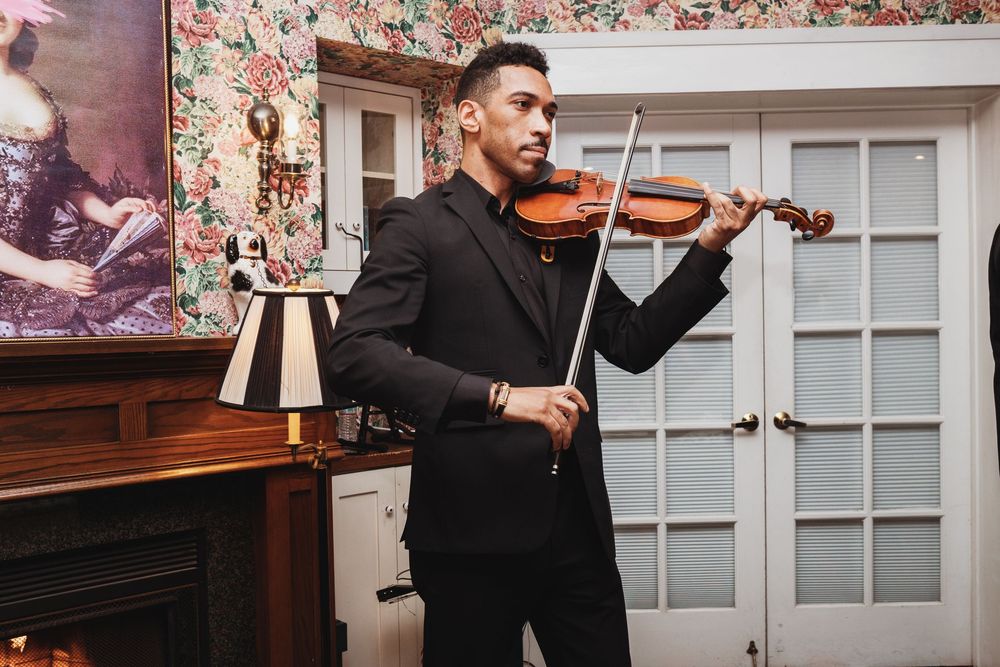 A man in a black suit plays violin in an elegant room with floral wallpaper and a fireplace.